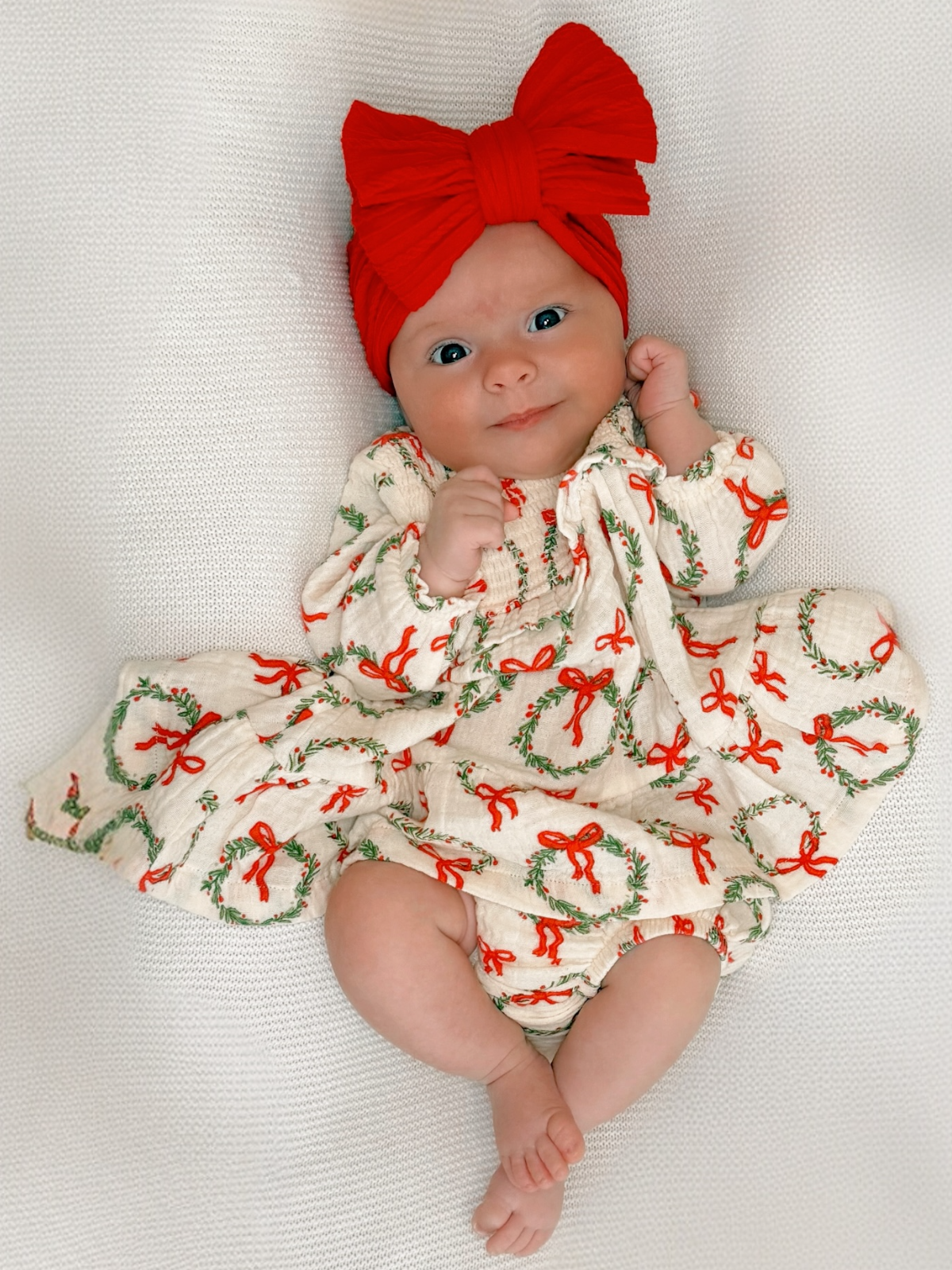 Baby girl in festive dress with red bows, smiling and wearing a large red headband, posed on a white blanket.