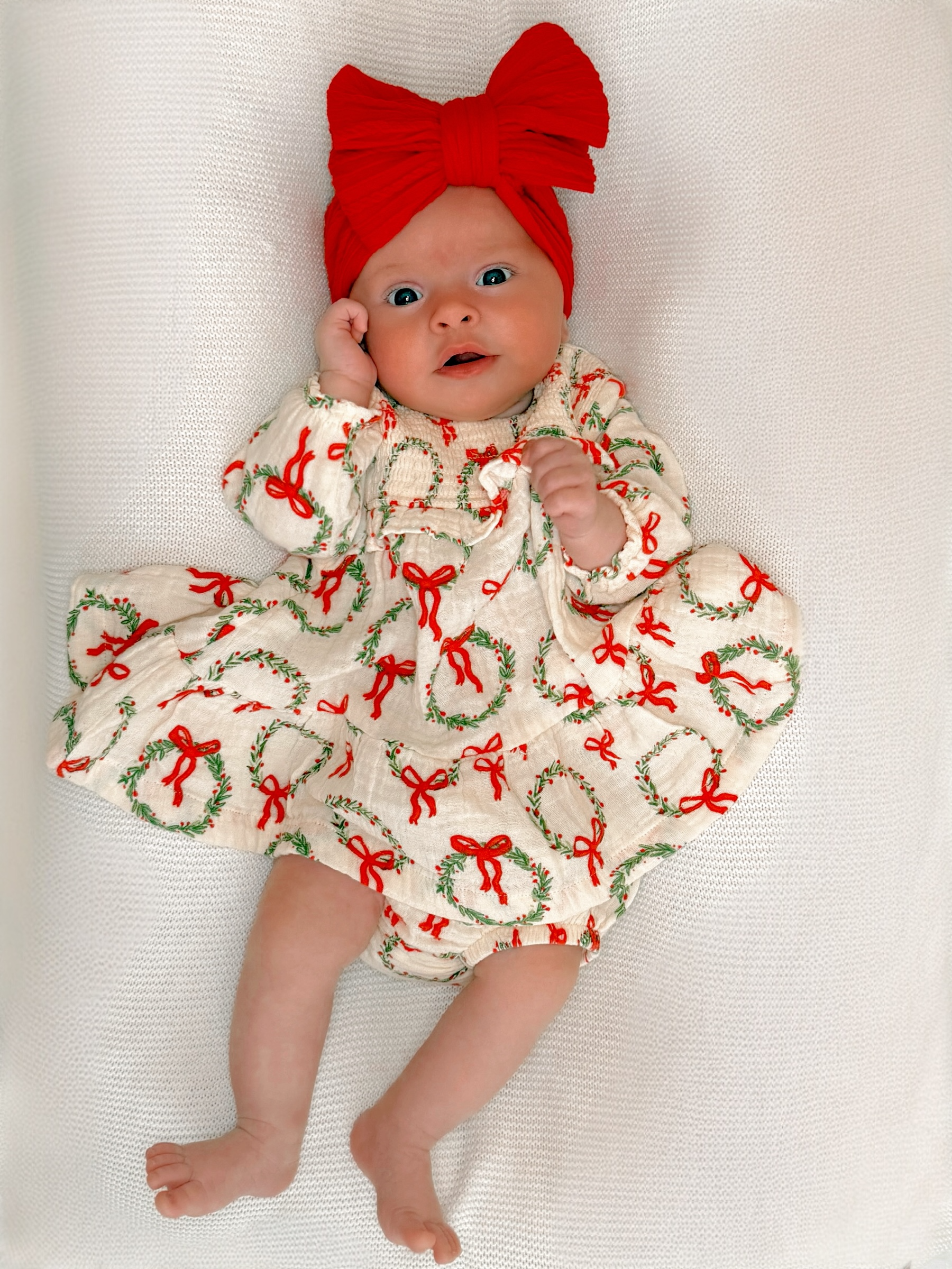 Baby dressed in a festive outfit with red bows and a large matching headband, lying on a soft white surface.