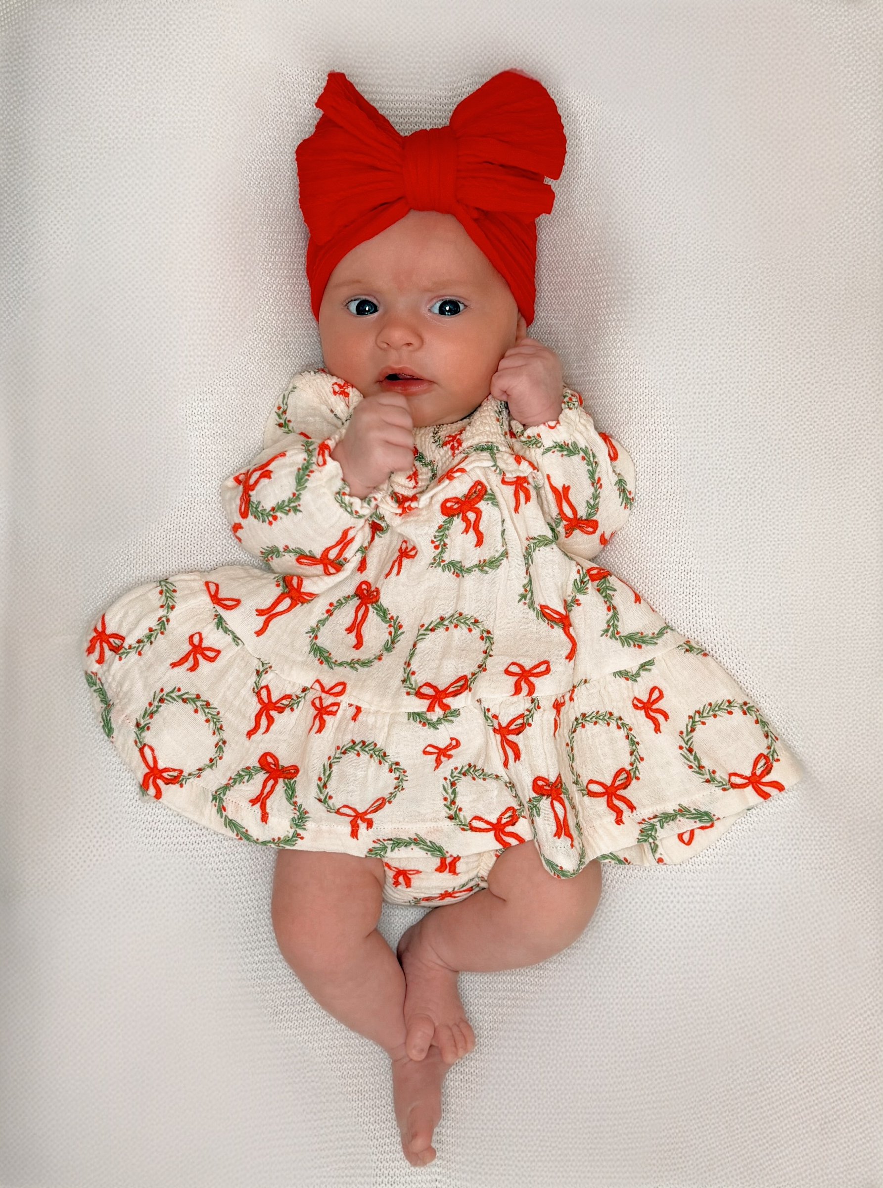 Newborn baby in a festive dress with holiday wreaths and a large red bow headband, lying on a textured surface.