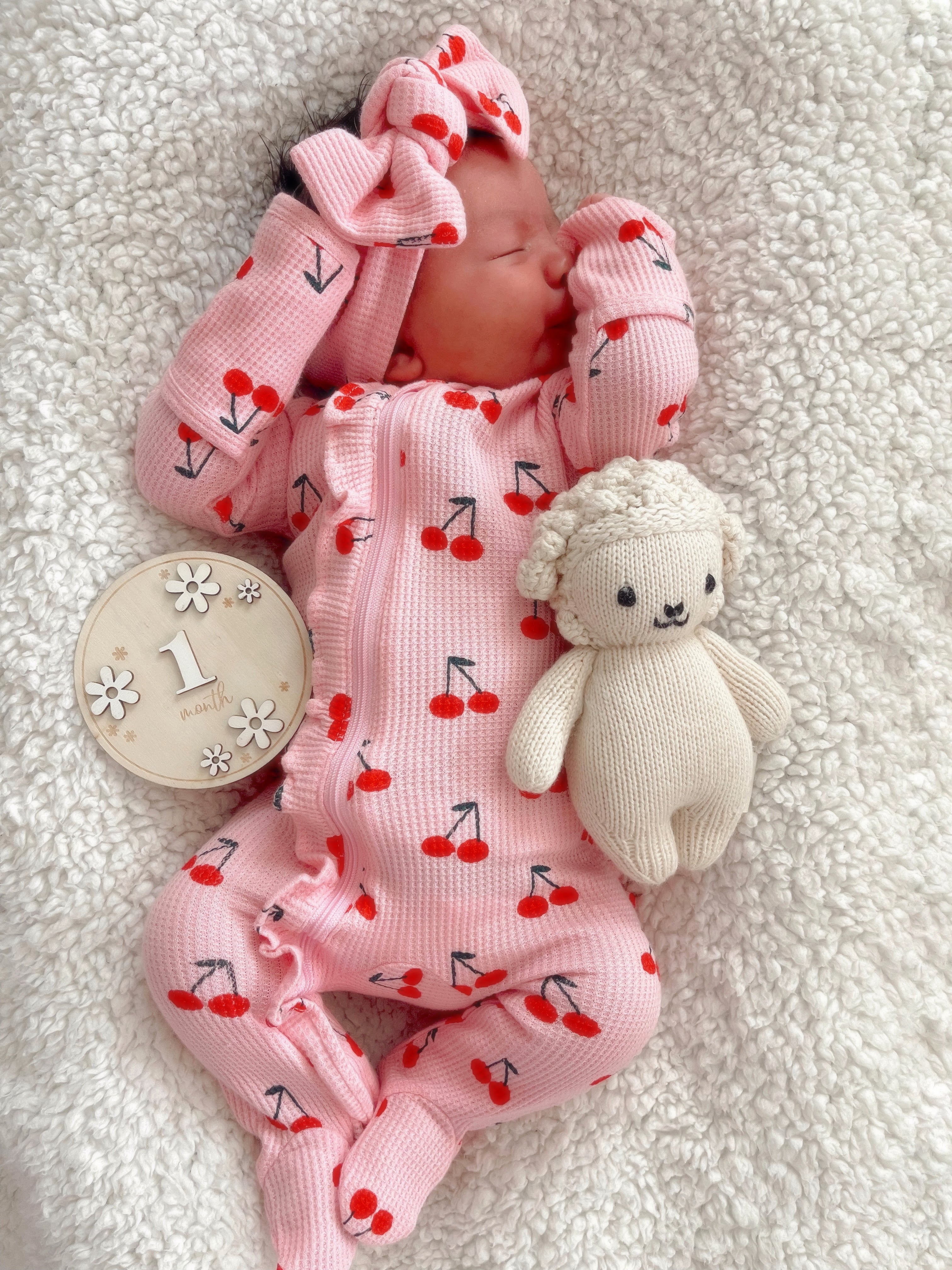 Newborn in cherry-patterned outfit, resting on a soft blanket with a plush toy and milestone marker.