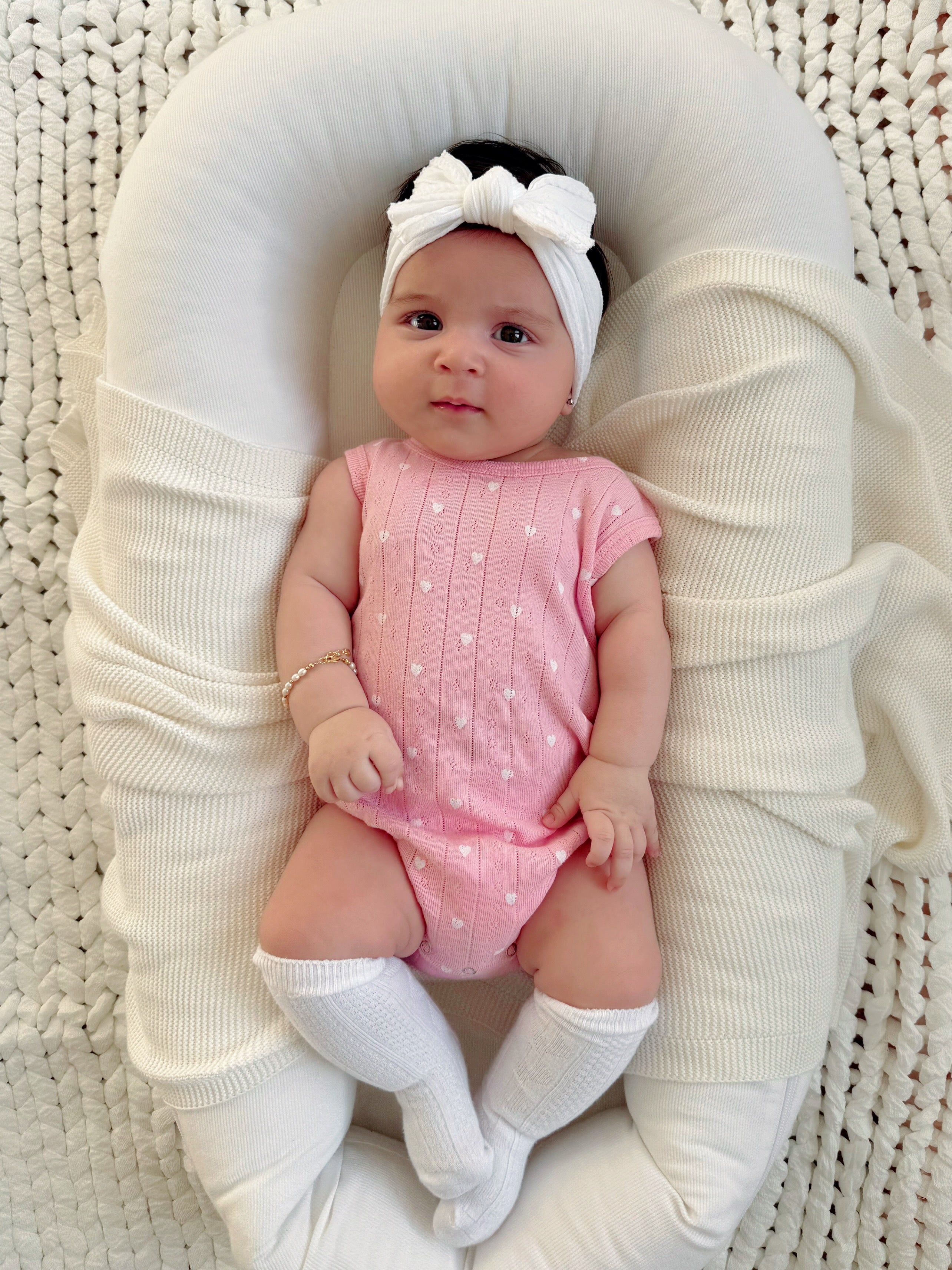 Baby girl in a pink outfit and headband, resting on a soft blanket with a cozy, textured background.