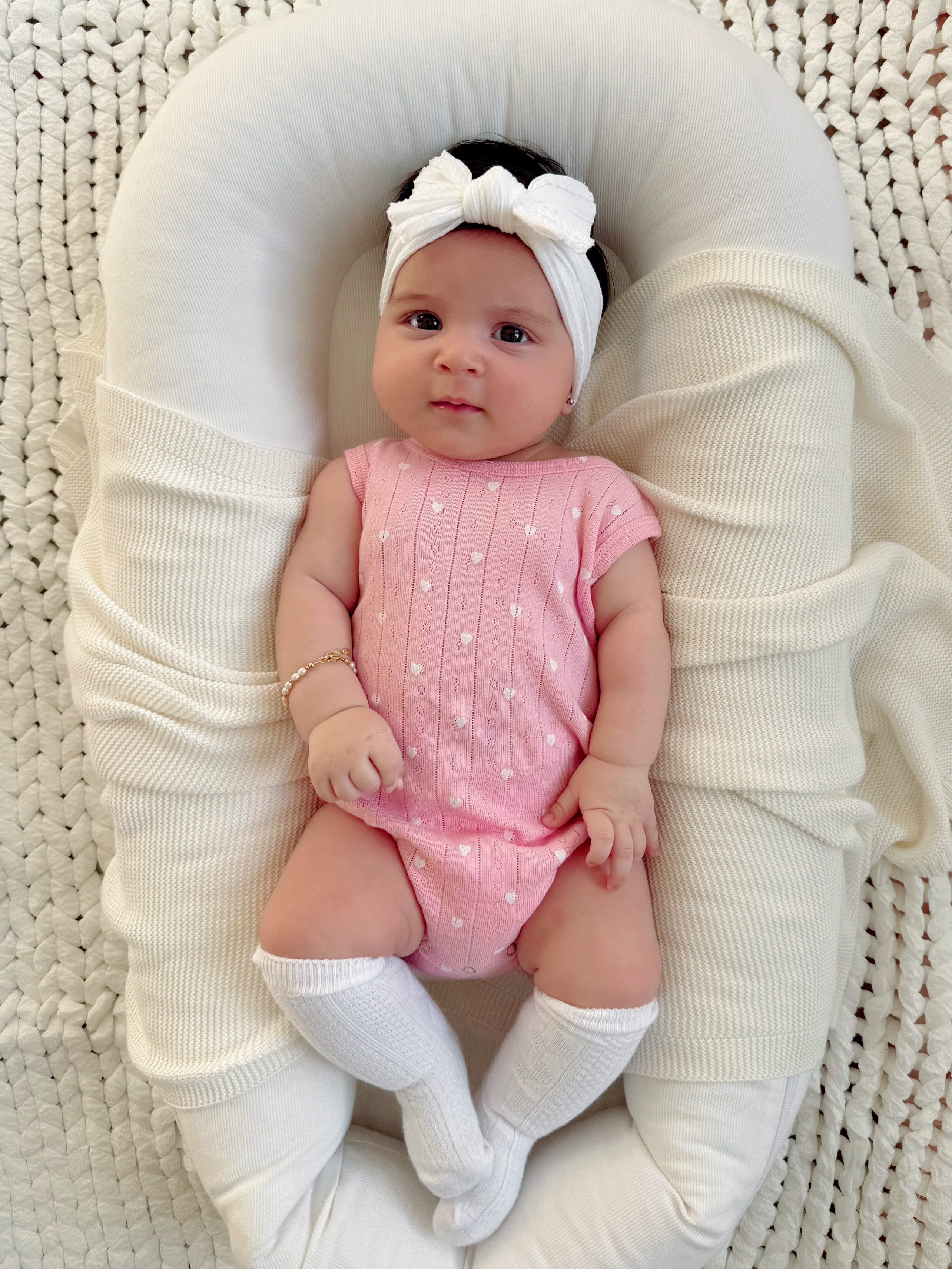 Baby girl in a pink outfit and headband, resting on a soft blanket with a cozy, textured background.