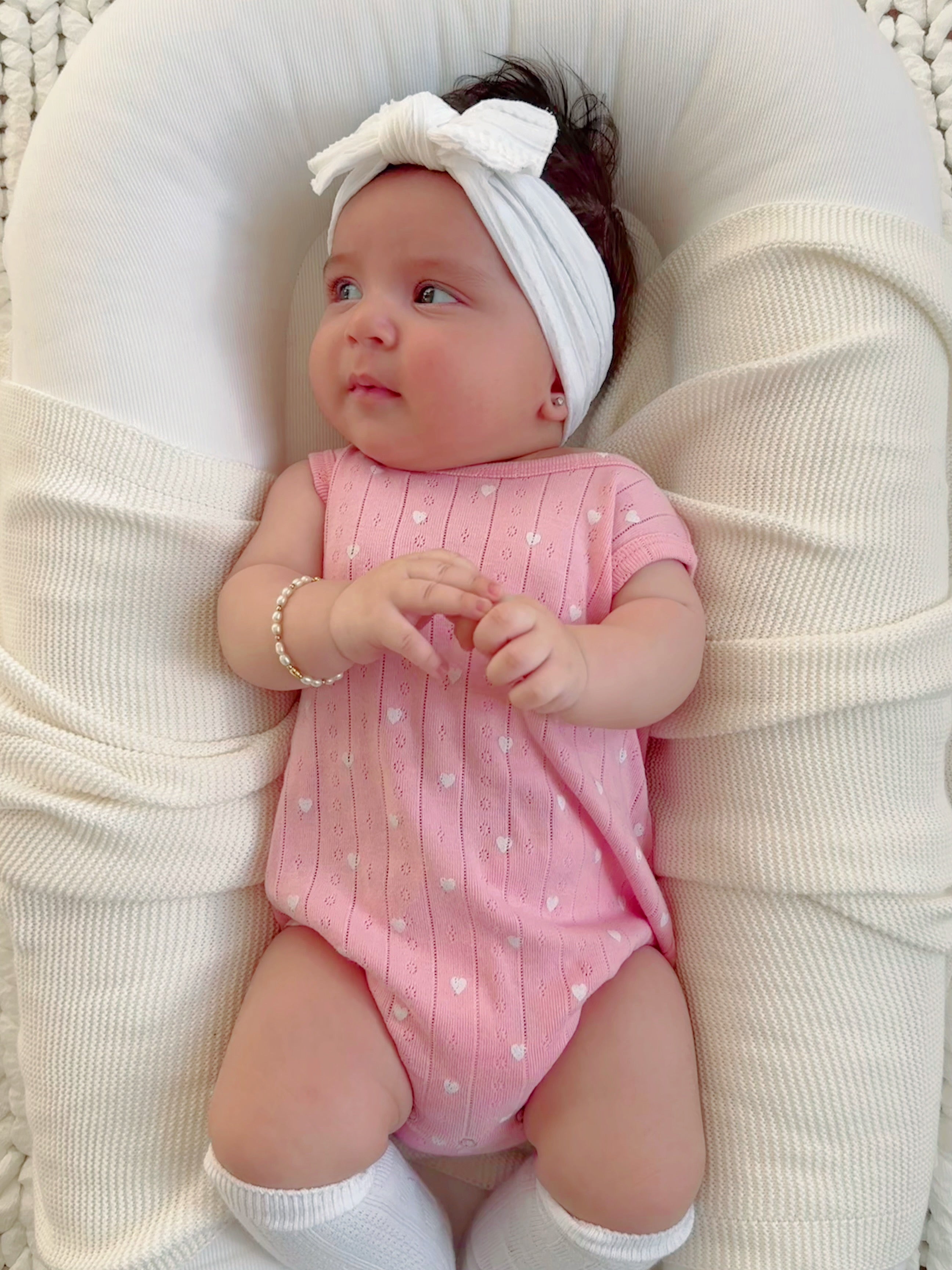Baby in a pink outfit with a white headband, sitting on a soft, textured surface, looking thoughtfully to the side.
