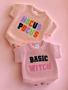 Two pink baby outfits with graphic text: "Hocus Pocus" on top and "Basic Witch" on the bottom, set against a pink background.