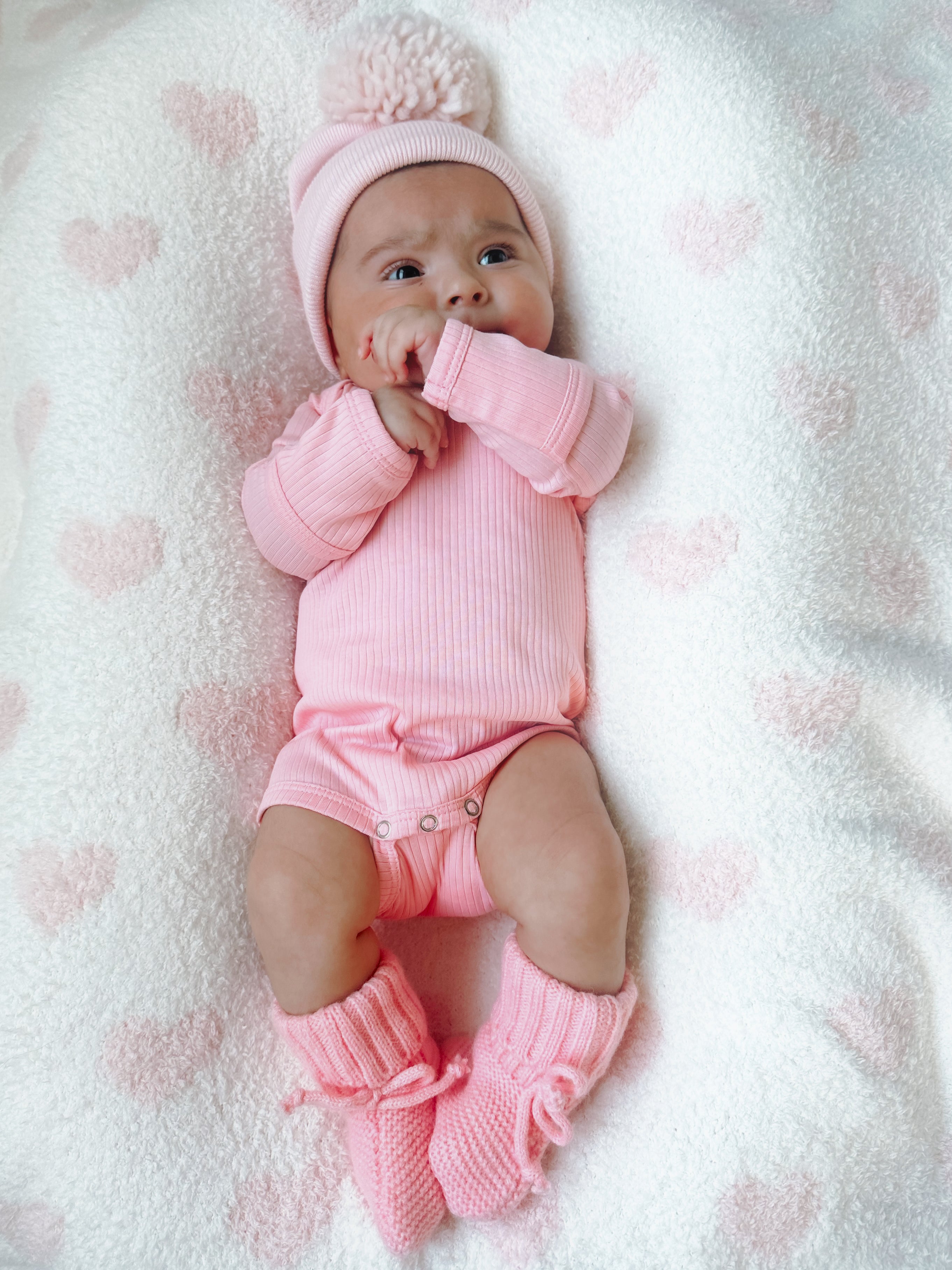 Baby girl in pink outfit with pom-pom hat lying on a soft, heart-patterned blanket, looking curiously at the camera.