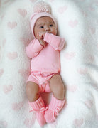Baby girl in pink outfit with pom-pom hat lying on a soft, heart-patterned blanket, looking curiously at the camera.