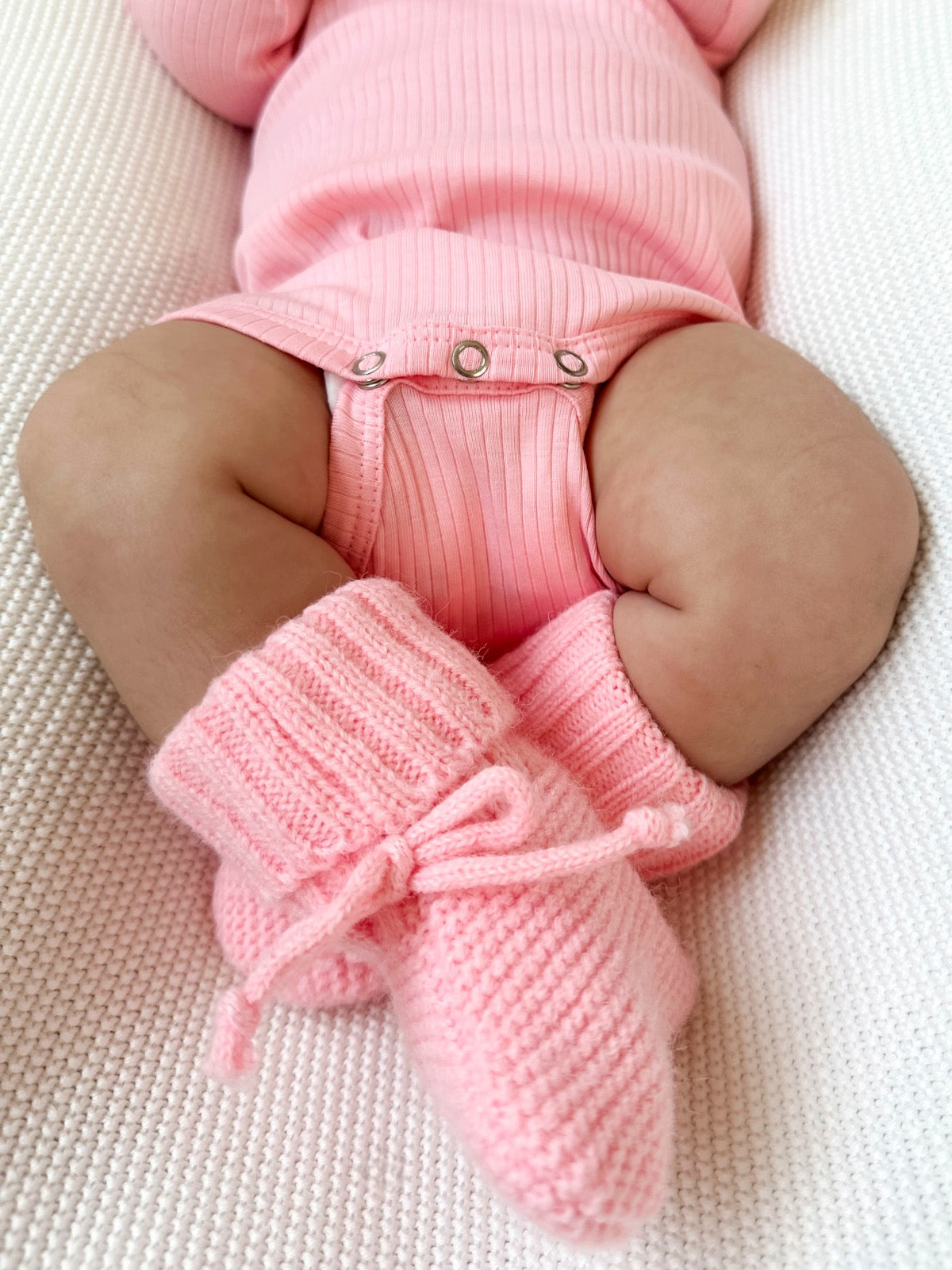 Baby legs in pink knitted booties, dressed in a ribbed pink onesie, laying on a textured white surface.