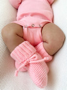 Baby legs in pink knitted booties, dressed in a ribbed pink onesie, laying on a textured white surface.