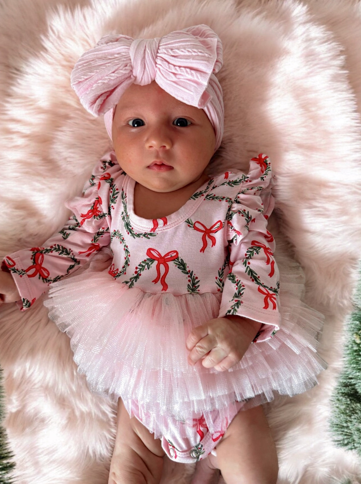 Baby girl in a pink bow and festive outfit, laying on a fluffy background, with a sweet expression.