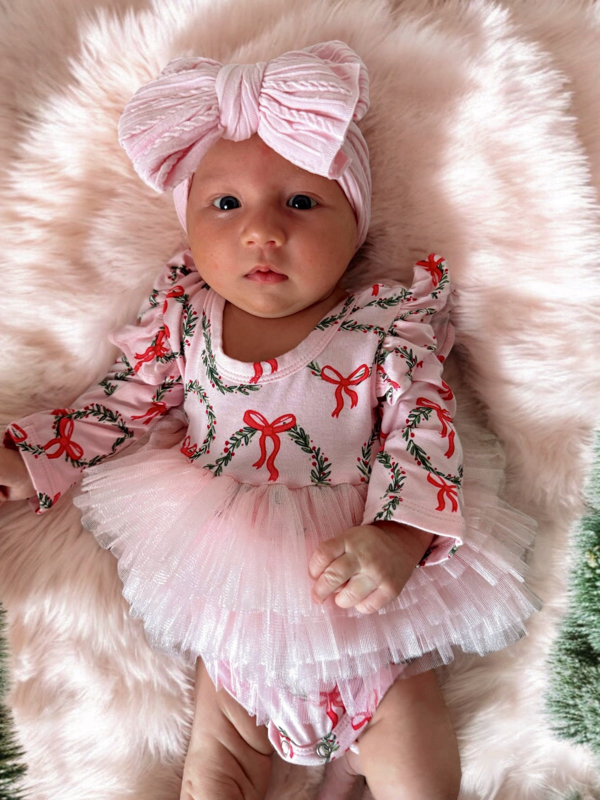 Baby girl in a pink bow and festive outfit, laying on a fluffy background, with a sweet expression.