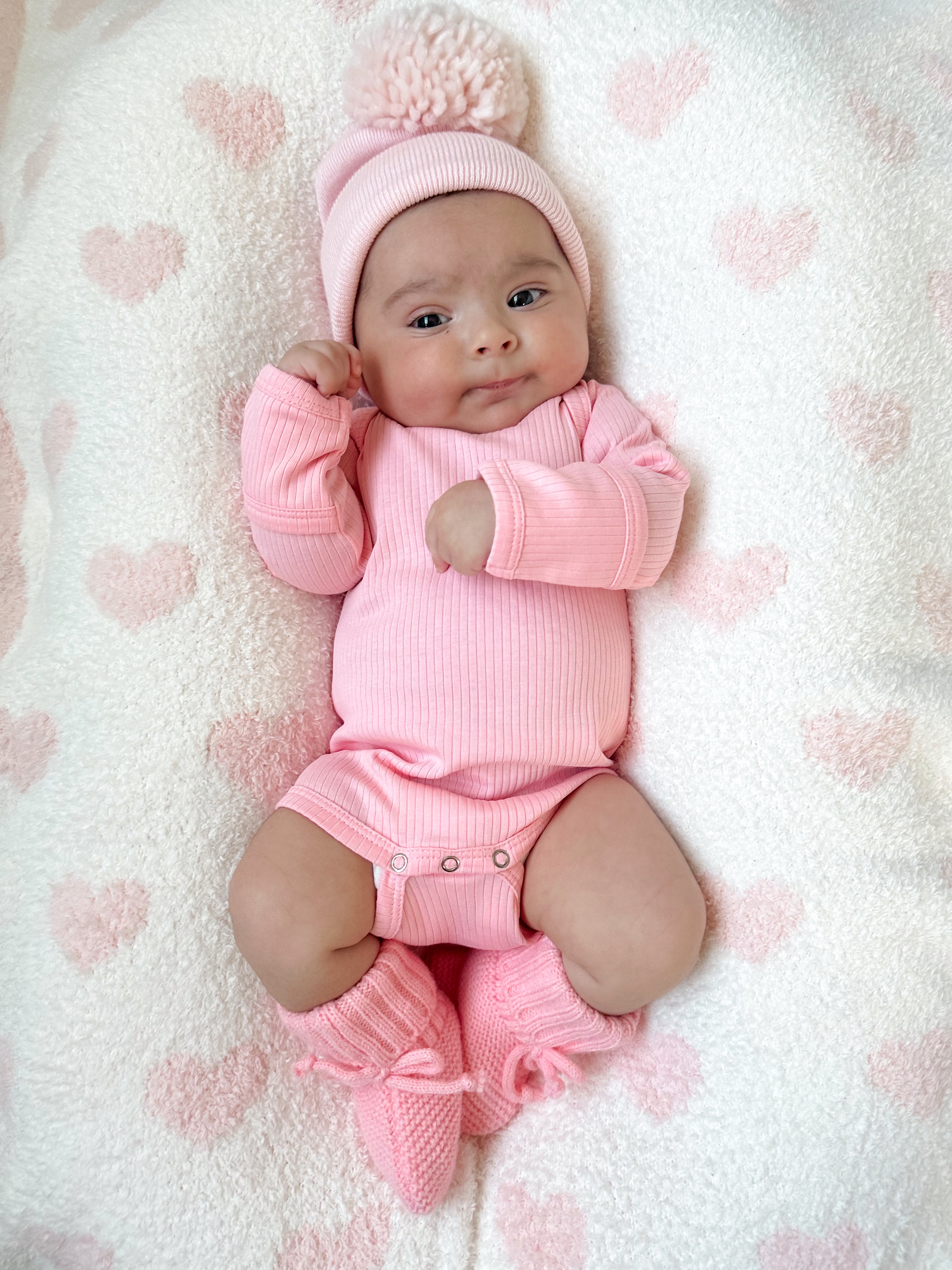 Adorable baby in pink attire and hat, lying on a soft, heart-patterned blanket.