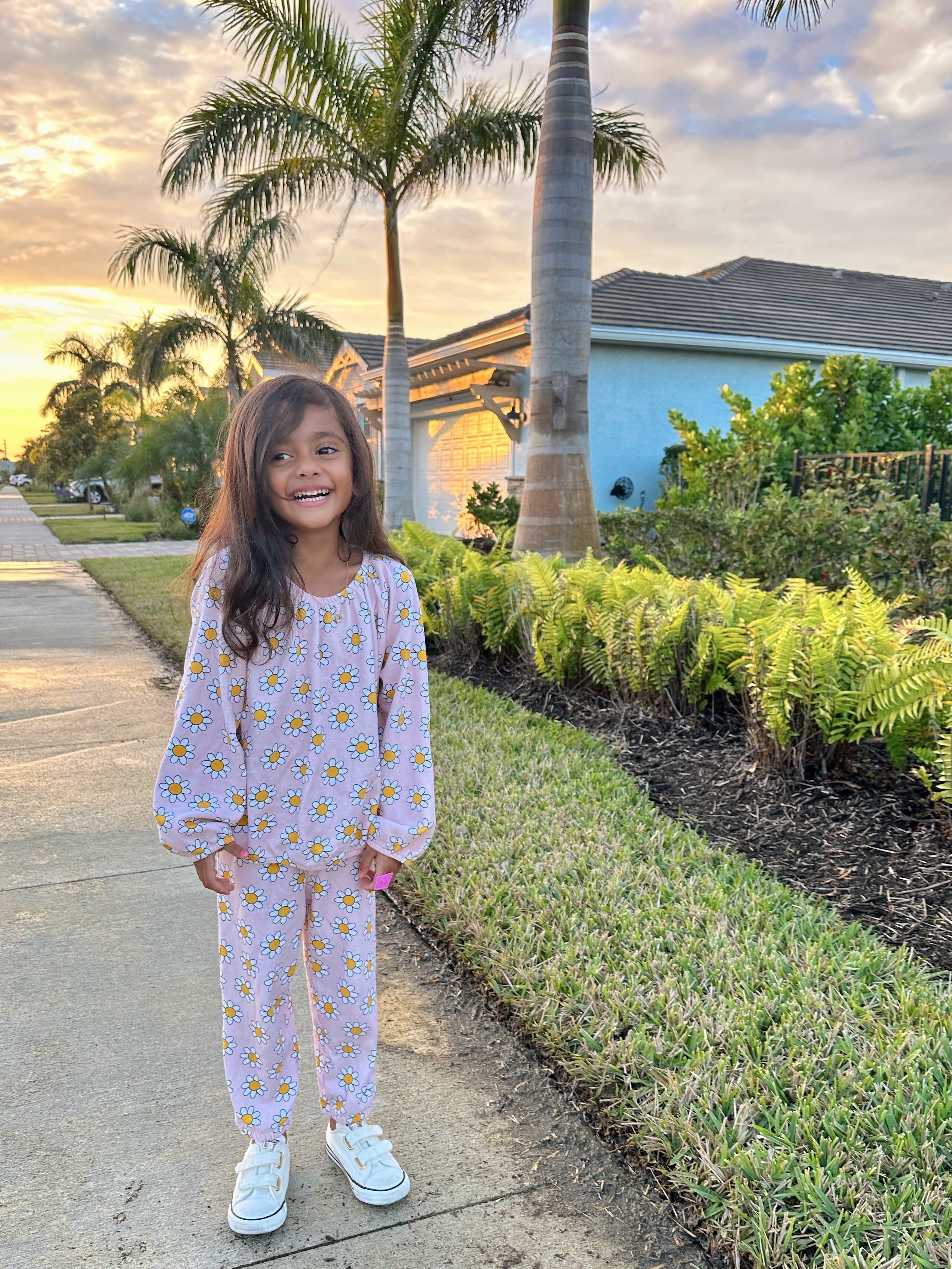 Smiling child in floral pajamas stands on a sidewalk with palm trees and a sunset backdrop.