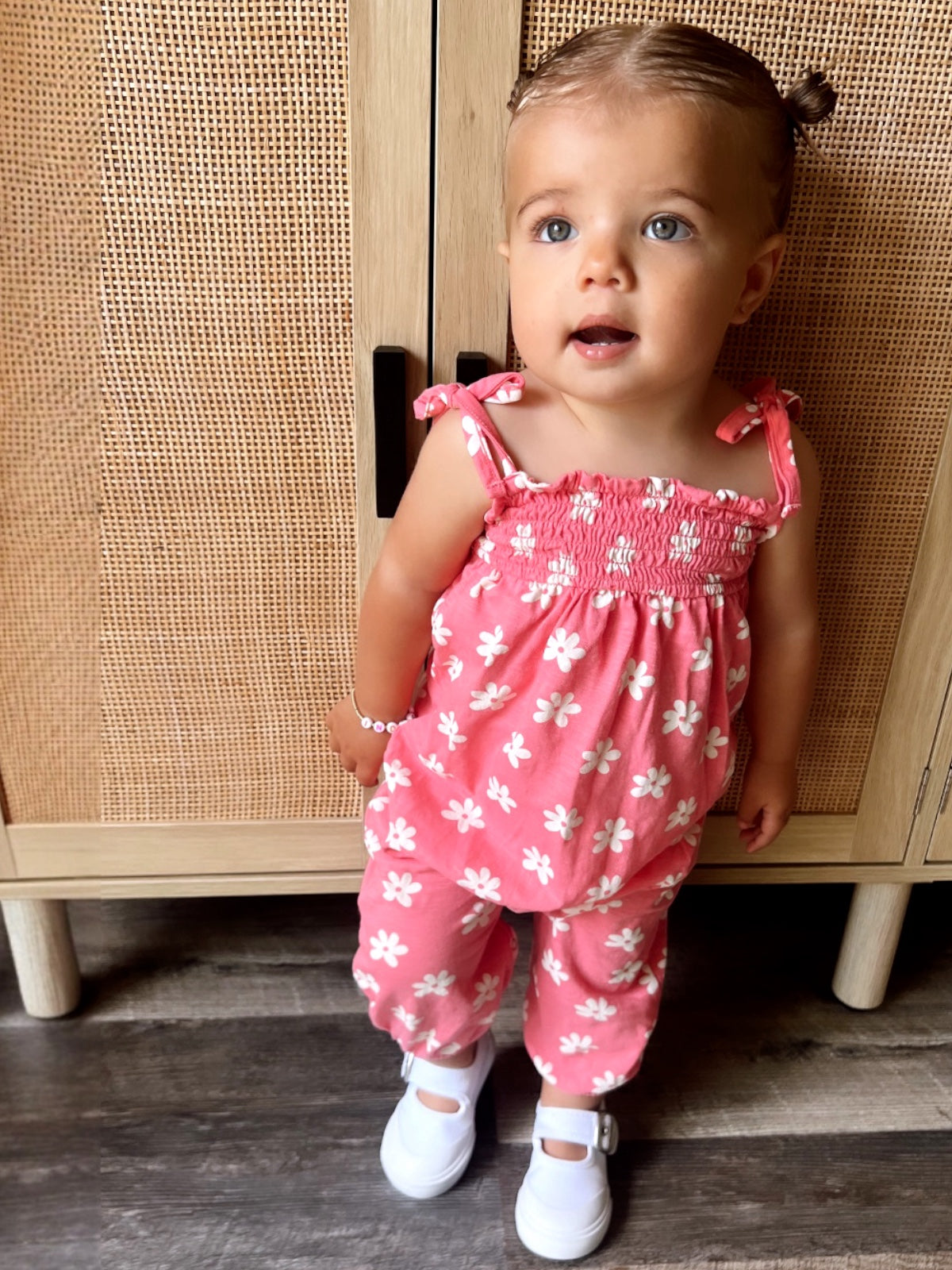 Toddler in a pink floral outfit stands in front of a wooden cabinet, wearing white shoes and a charming expression.