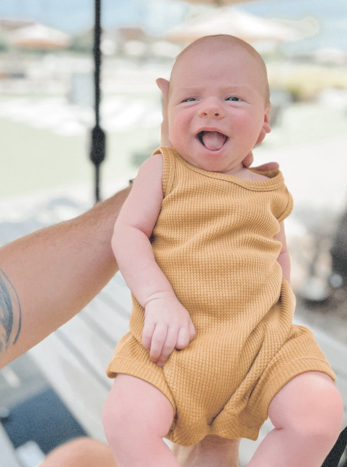 Smiling baby in a yellow outfit being held up. Soft outdoor background with blurred umbrellas.