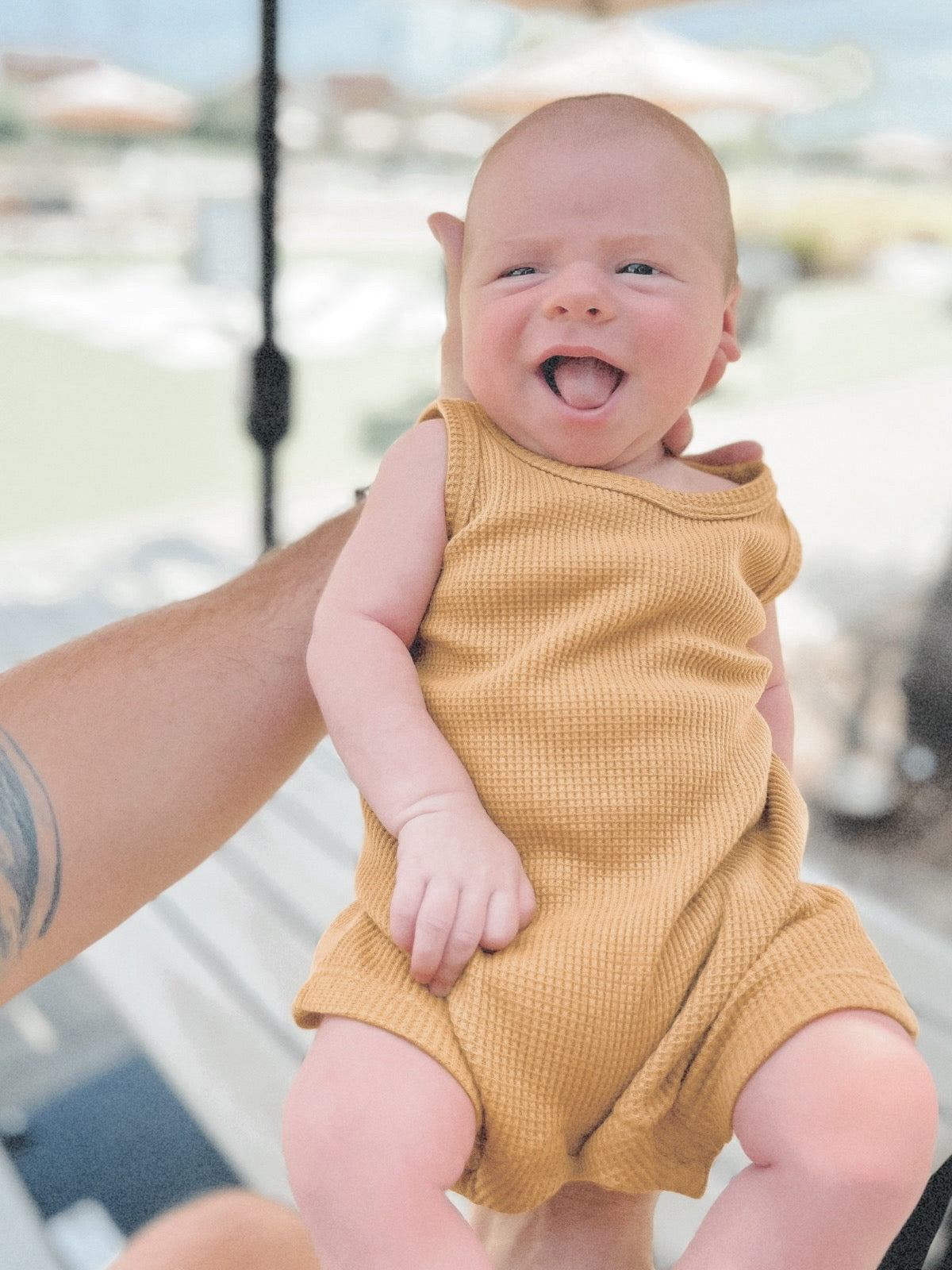 Smiling baby in a yellow outfit being held up. Soft outdoor background with blurred umbrellas.
