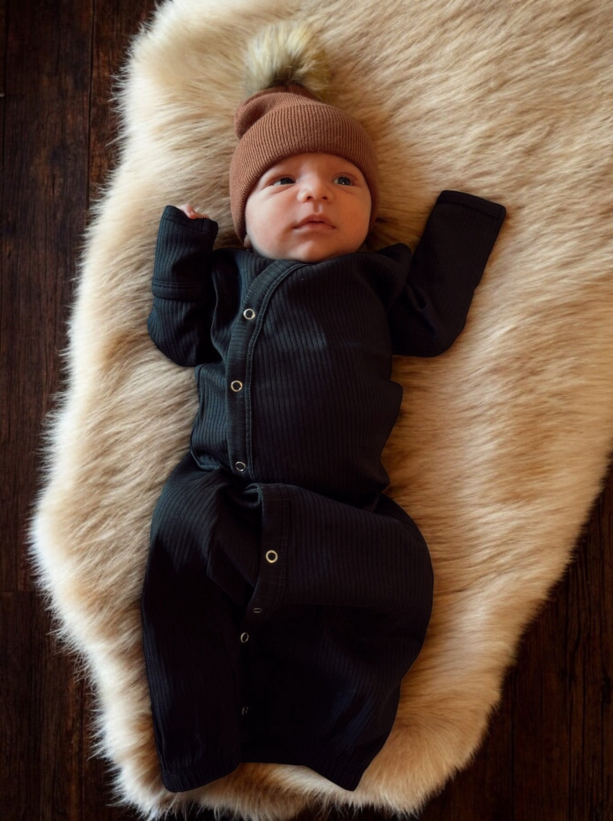 Newborn in a black onesie and brown beanie, lying on a fluffy cream blanket.