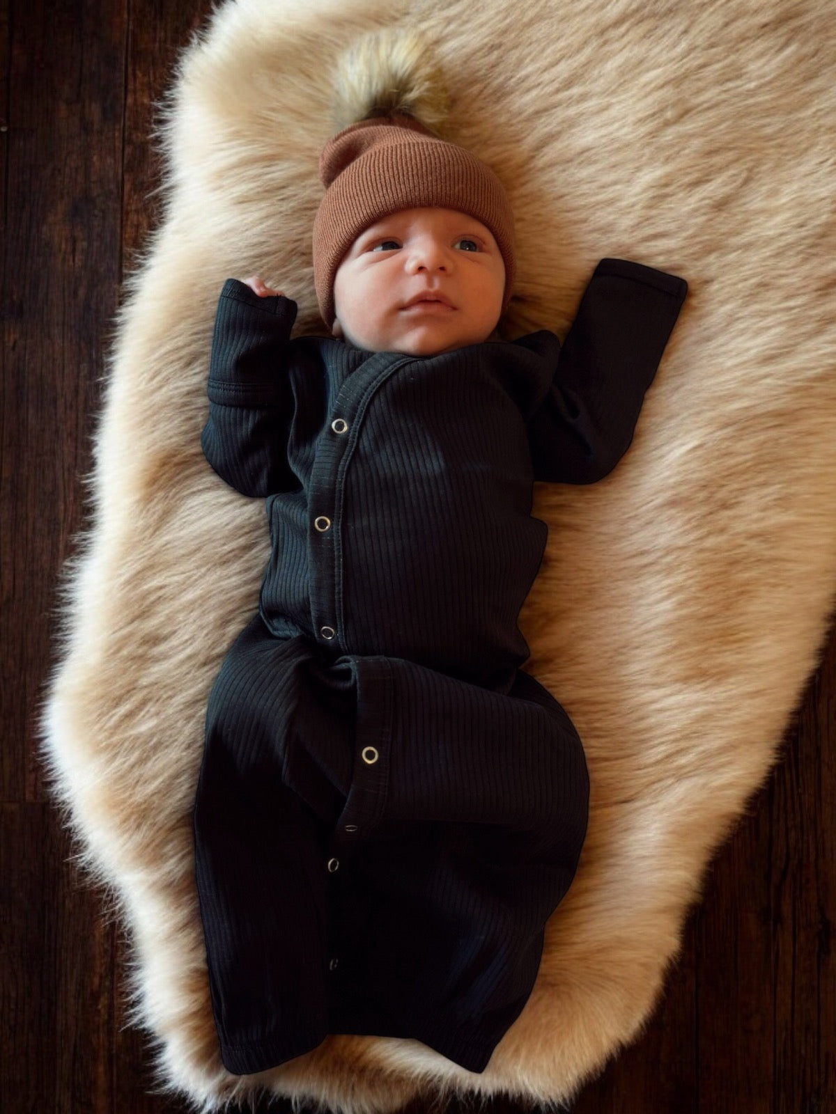 Newborn in a black onesie and brown beanie, lying on a fluffy cream blanket.