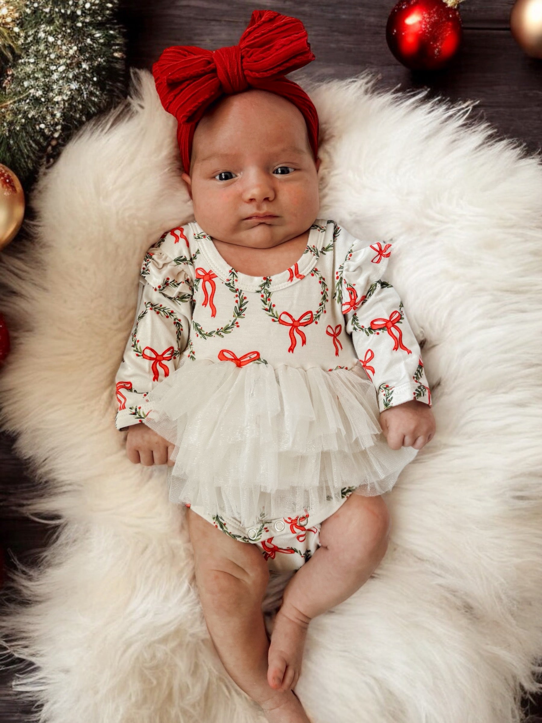 Baby girl in festive outfit with red bows, lying on soft white fur surrounded by holiday decorations.