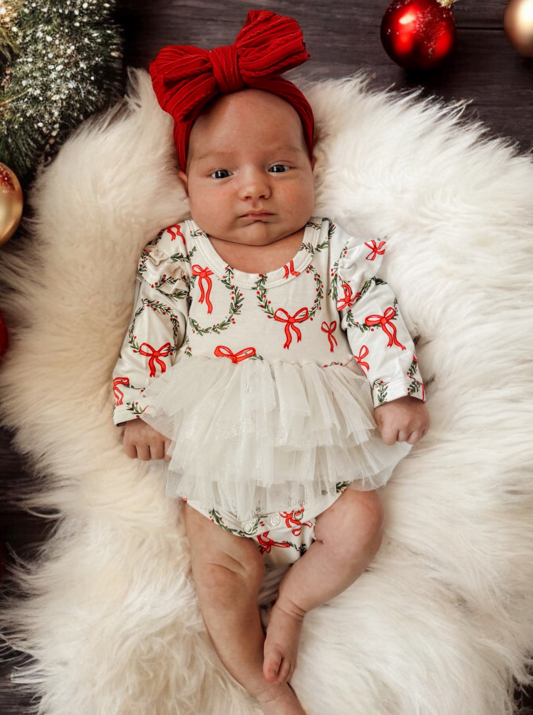 Baby girl in festive outfit with red bows, lying on soft white fur surrounded by holiday decorations.