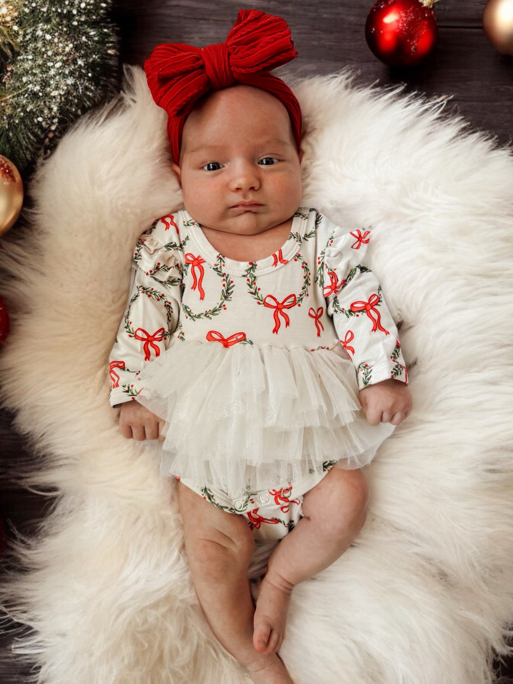 Baby girl in festive outfit with red bows, lying on soft white fur surrounded by holiday decorations.