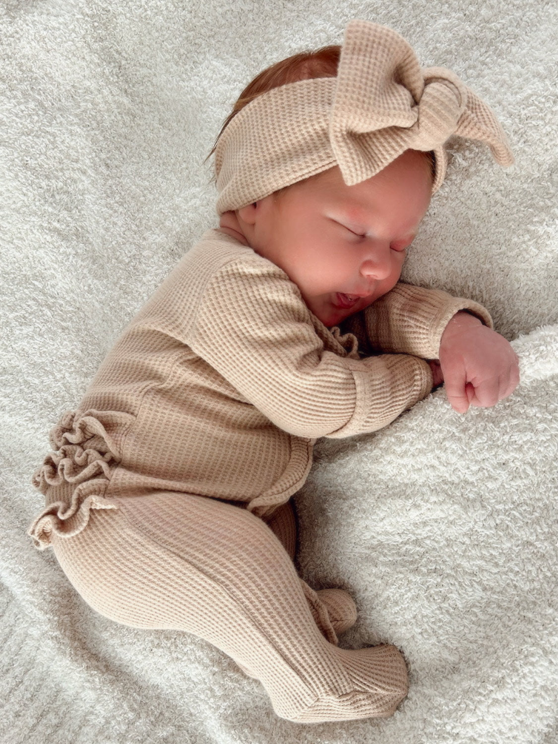 Sleeping baby in a beige outfit and headband, resting on a soft, textured blanket.