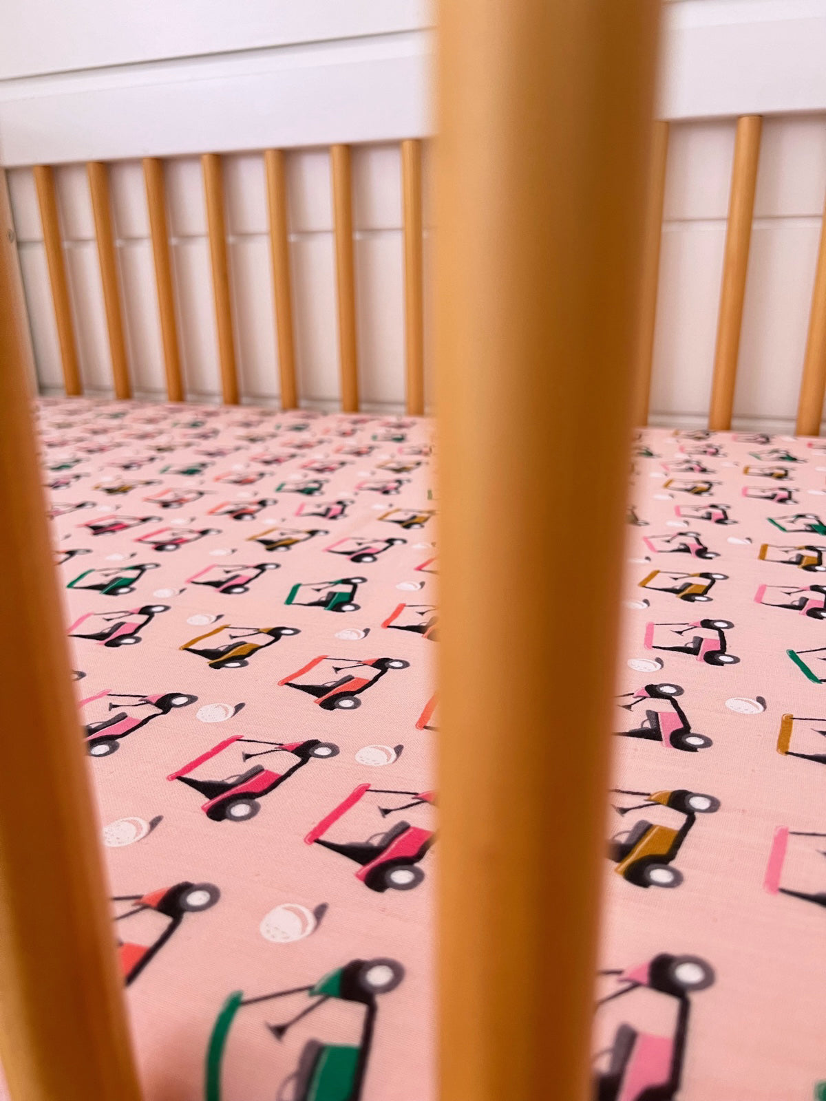 Baby crib with a patterned pink sheet featuring colorful toy cars and baseballs, viewed through wooden slats.