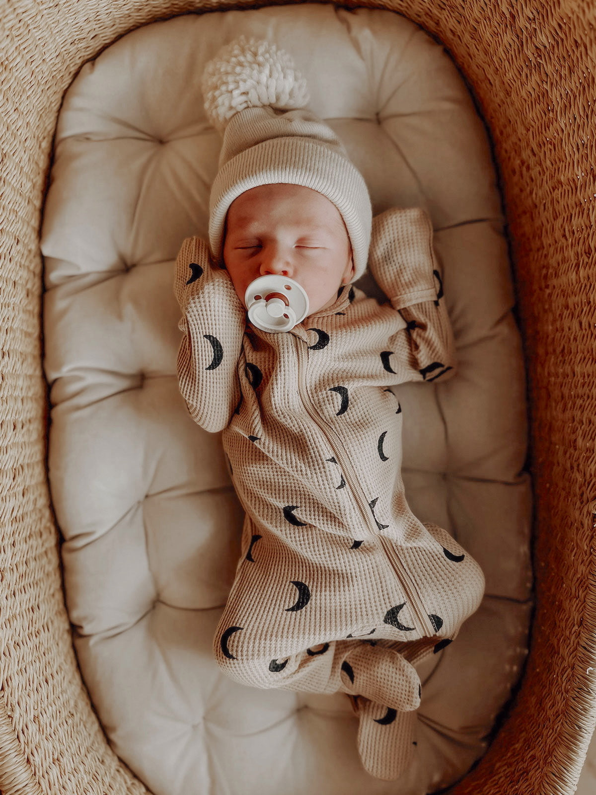 Baby peacefully sleeping in a cozy bassinet, wearing a patterned onesie and a soft pom-pom hat.