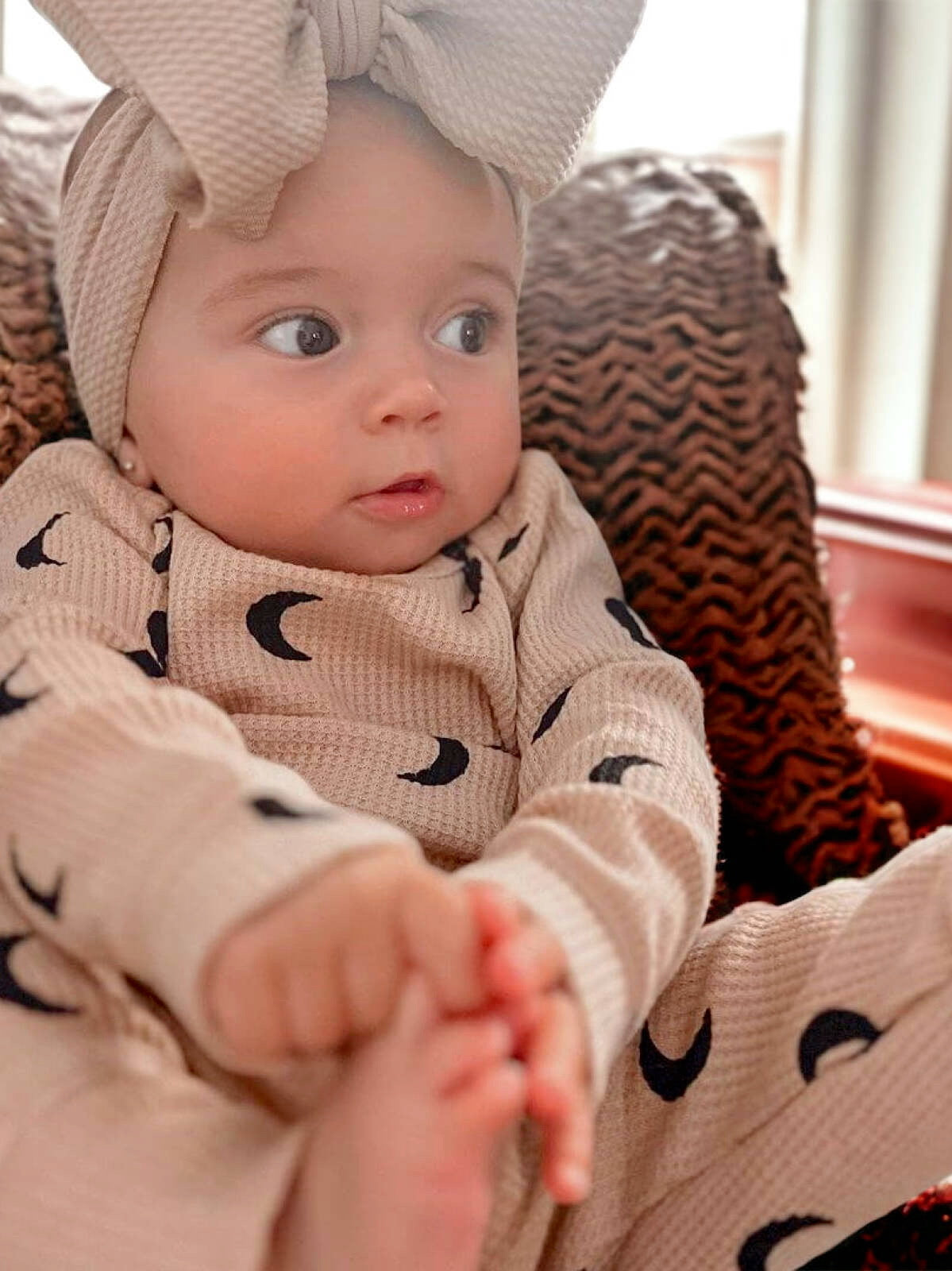 Baby with a big bow headband, wearing a patterned outfit, sitting comfortably and playing with their feet.