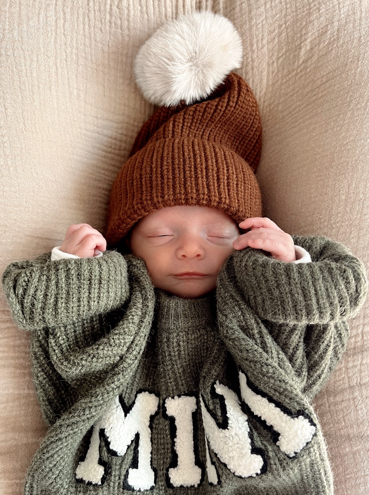 A sleeping baby in a cozy sweater and a brown knit hat with a pom-pom, resting on a soft beige blanket.