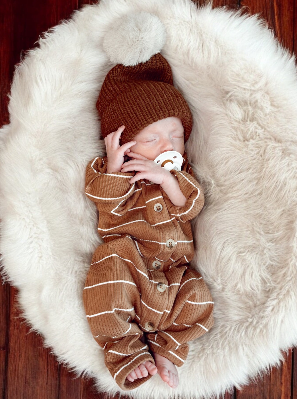 Sleeping newborn in a brown outfit with a pom-pom hat, resting on a fluffy white blanket.