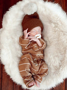 Sleeping newborn in a brown outfit with a pom-pom hat, resting on a fluffy white blanket.