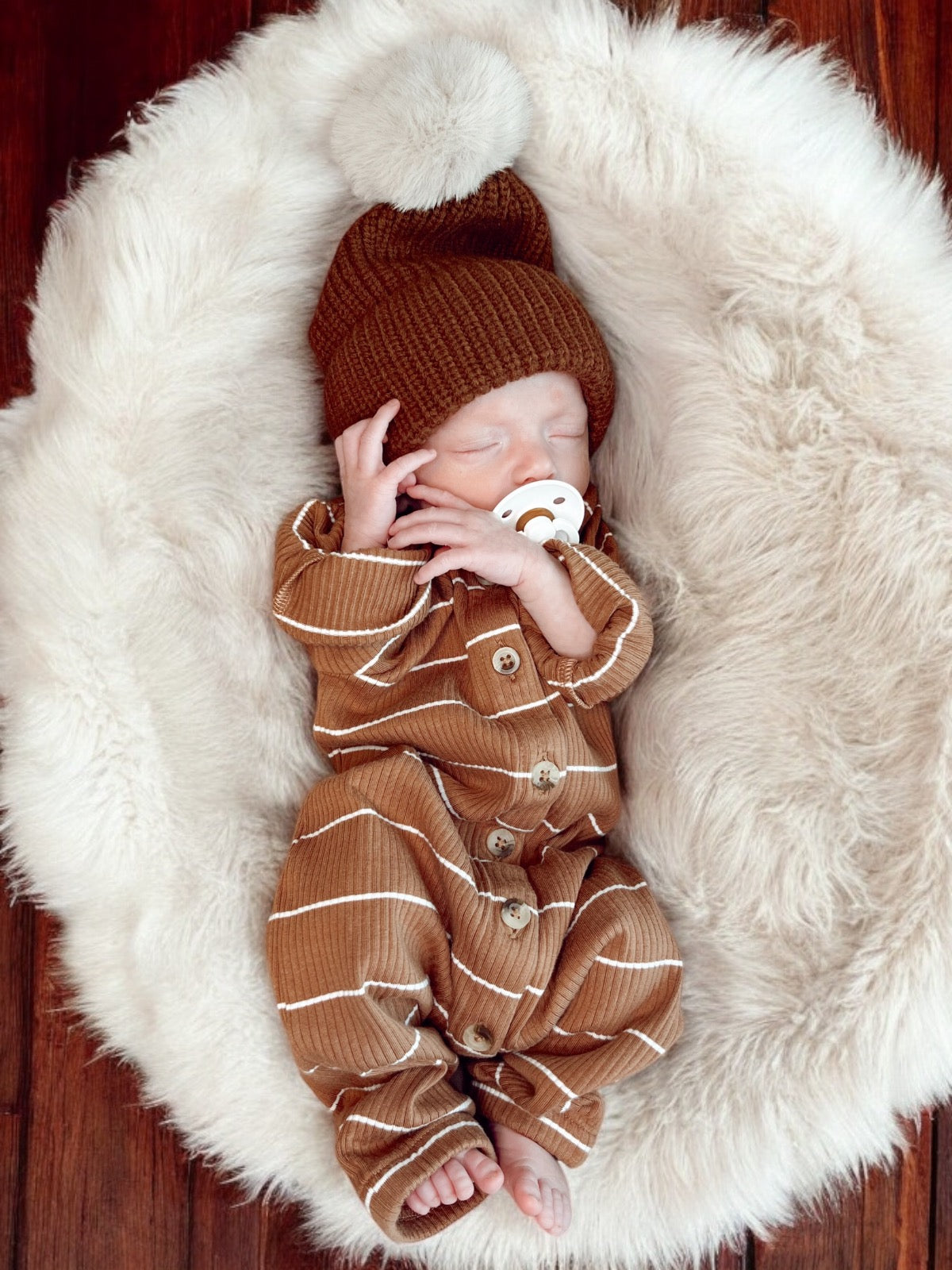 Sleeping newborn in a brown outfit with a pom-pom hat, resting on a fluffy white blanket.