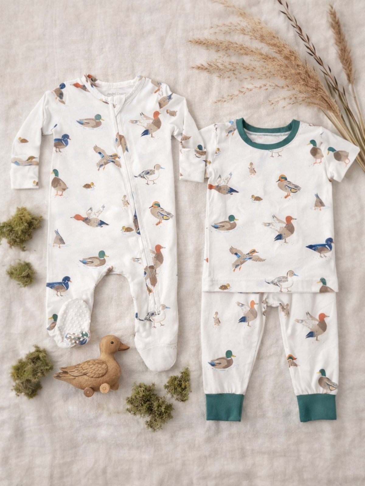 Two sets of children's pajamas featuring duck patterns, alongside a wooden duck toy and green moss accents.