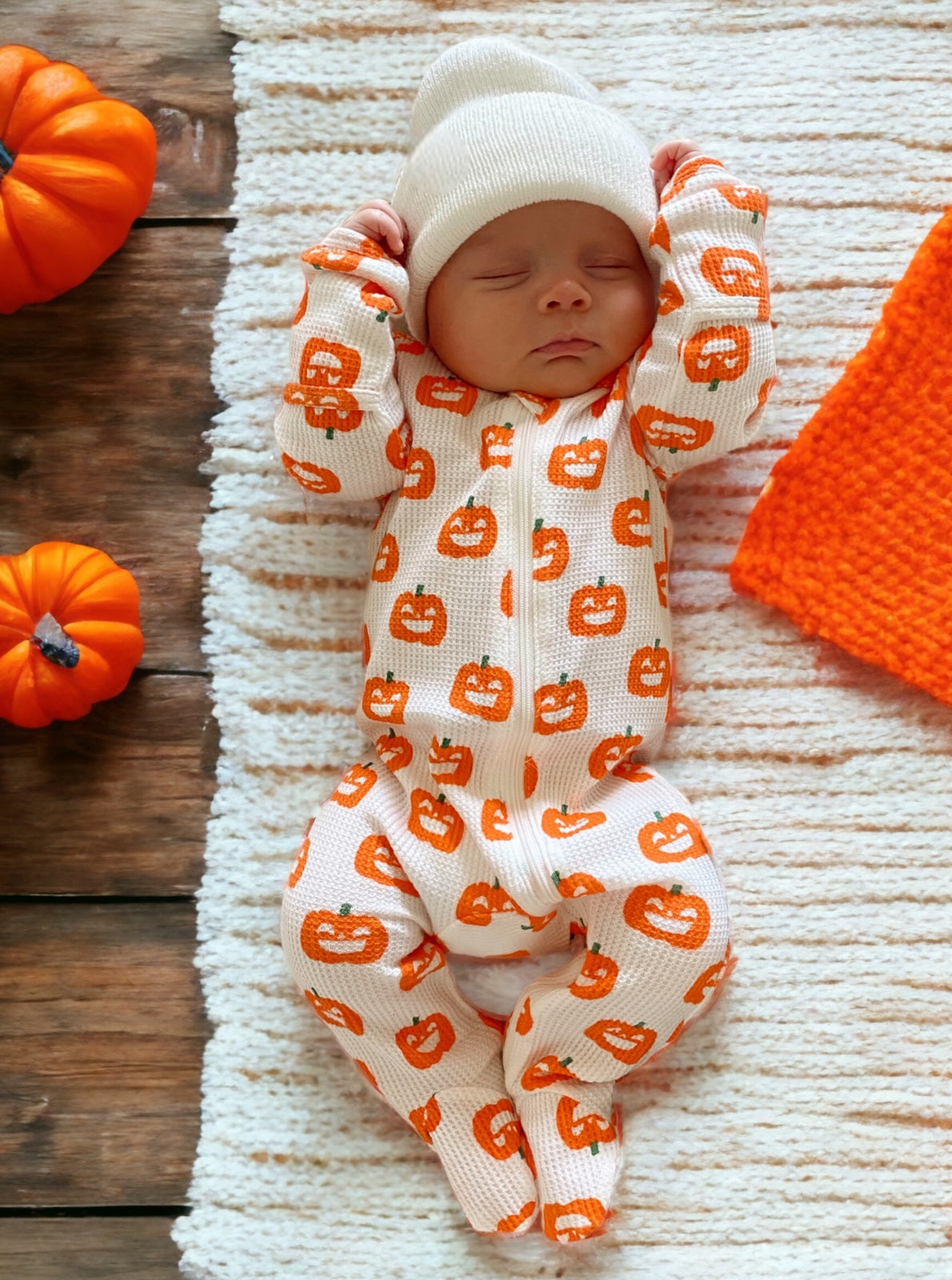 Newborn in pumpkin-patterned outfit, sleeping on a textured blanket, surrounded by small pumpkins and an orange cloth.