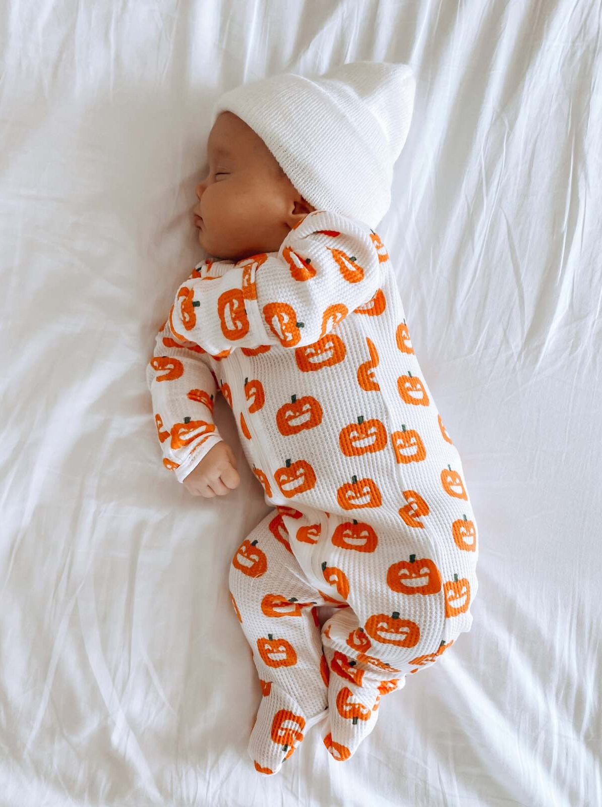 Sleeping baby in pumpkin-patterned outfit on a white blanket, wearing a white knit hat.