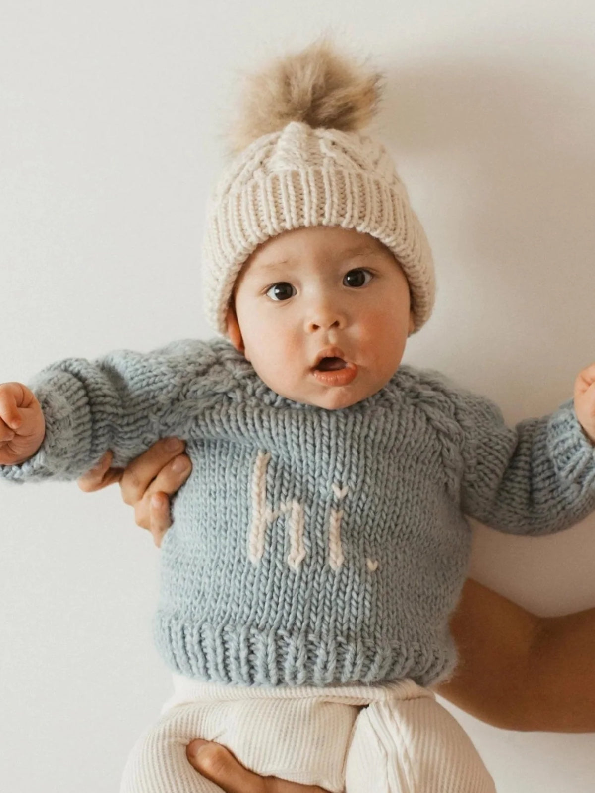 Baby in a blue sweater with "hi." text, wearing a cream knit hat, held by a hand against a light background.