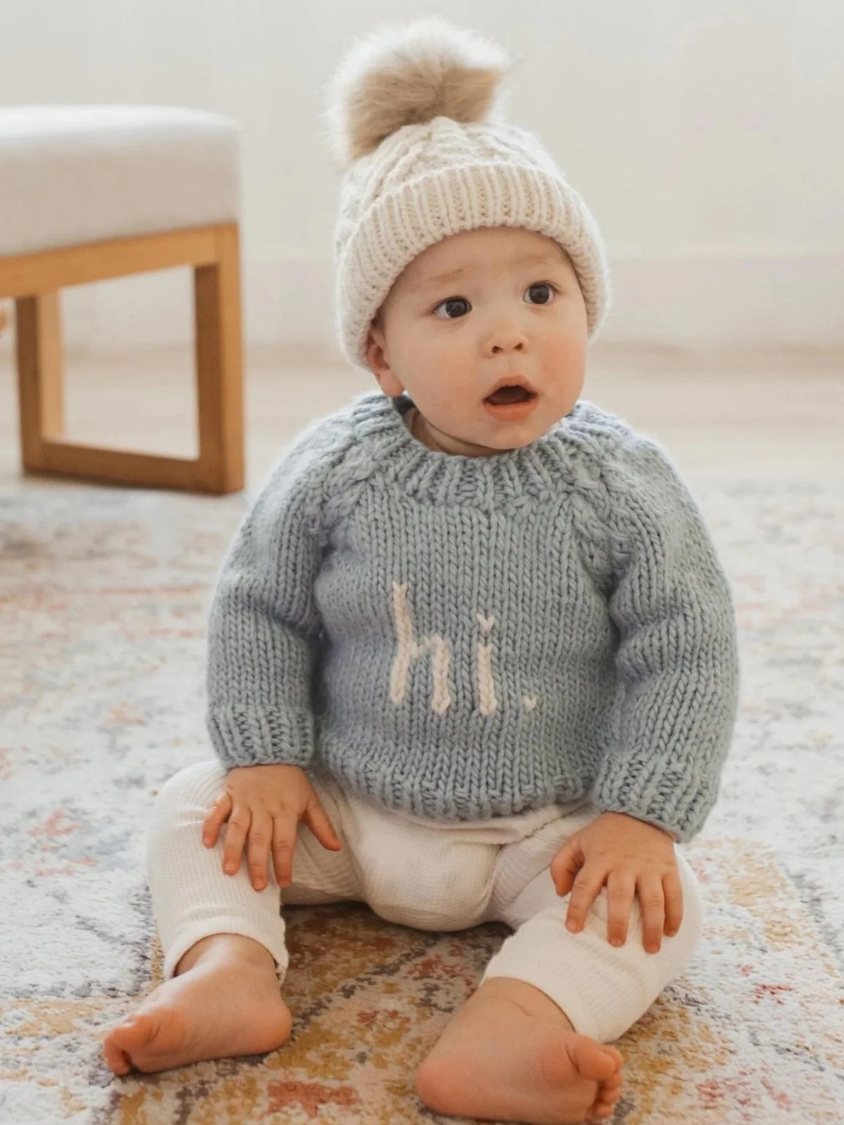 A surprised baby in a blue sweater and white pants sits on a rug, wearing a knit hat with a pom-pom.