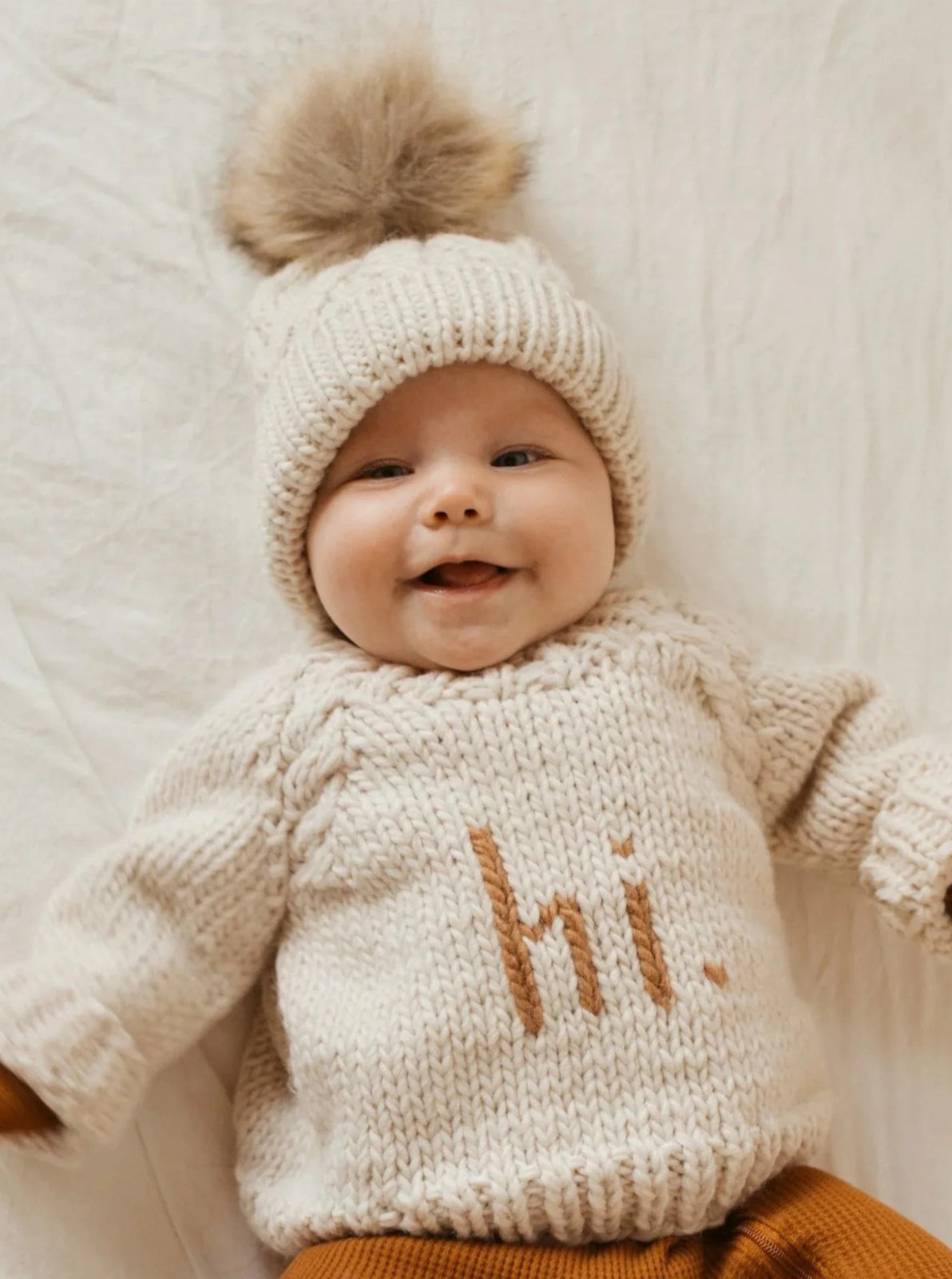 Smiling baby in cream sweater with "hi." text and winter hat with pom-pom, lying on a light-colored blanket.
