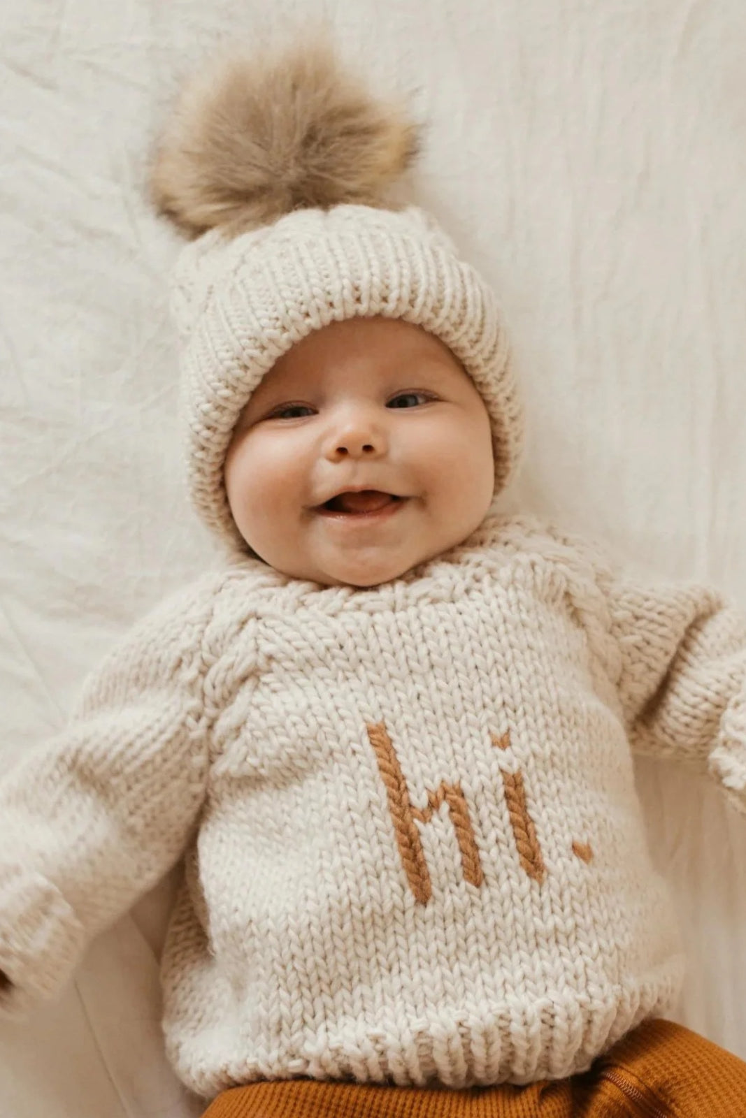 Smiling baby in cream sweater with "hi." text and winter hat with pom-pom, lying on a light-colored blanket.