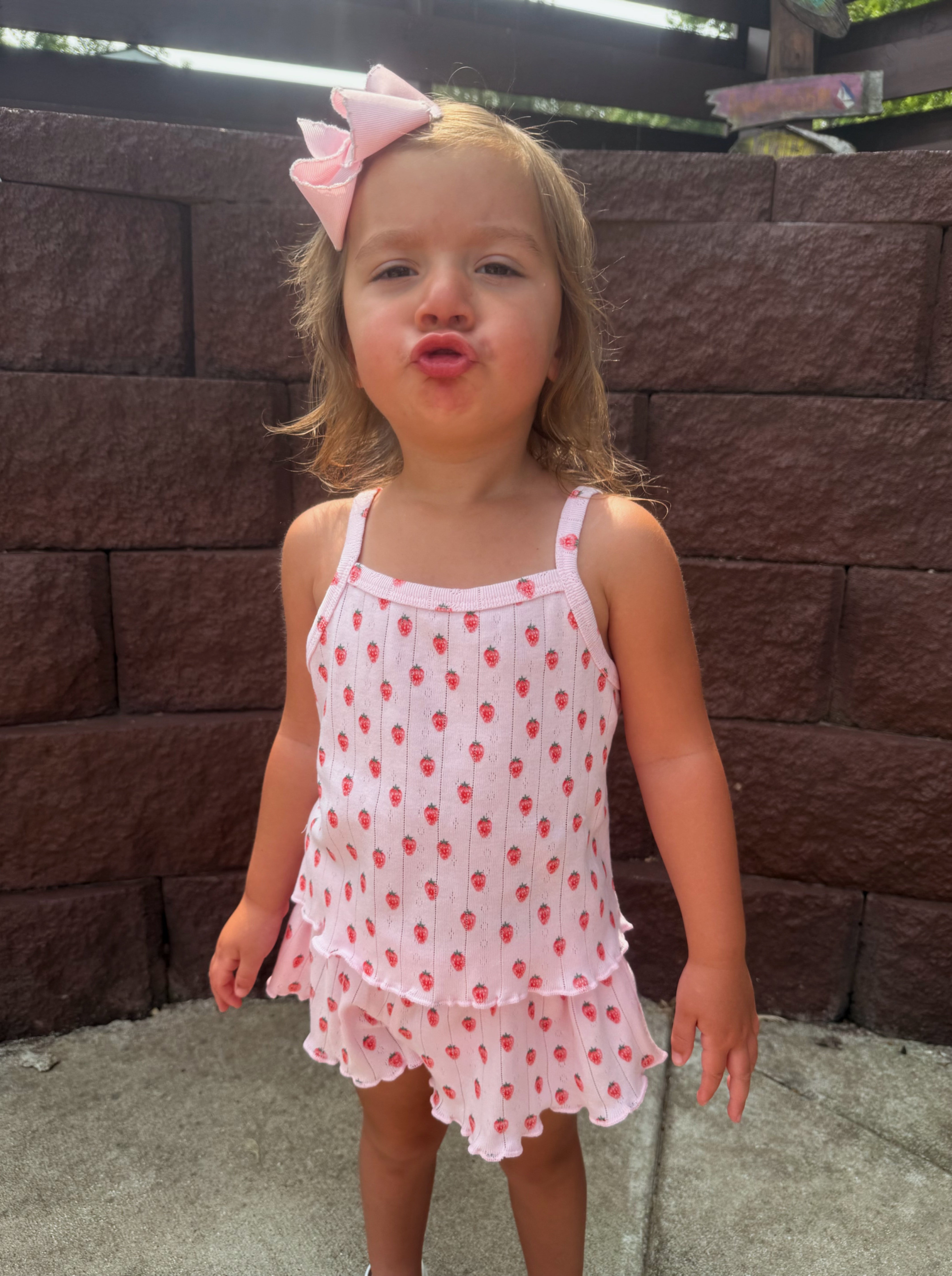 Young girl with a pink bow, wearing a strawberry-patterned outfit, making a playful face outdoors by a stone wall.