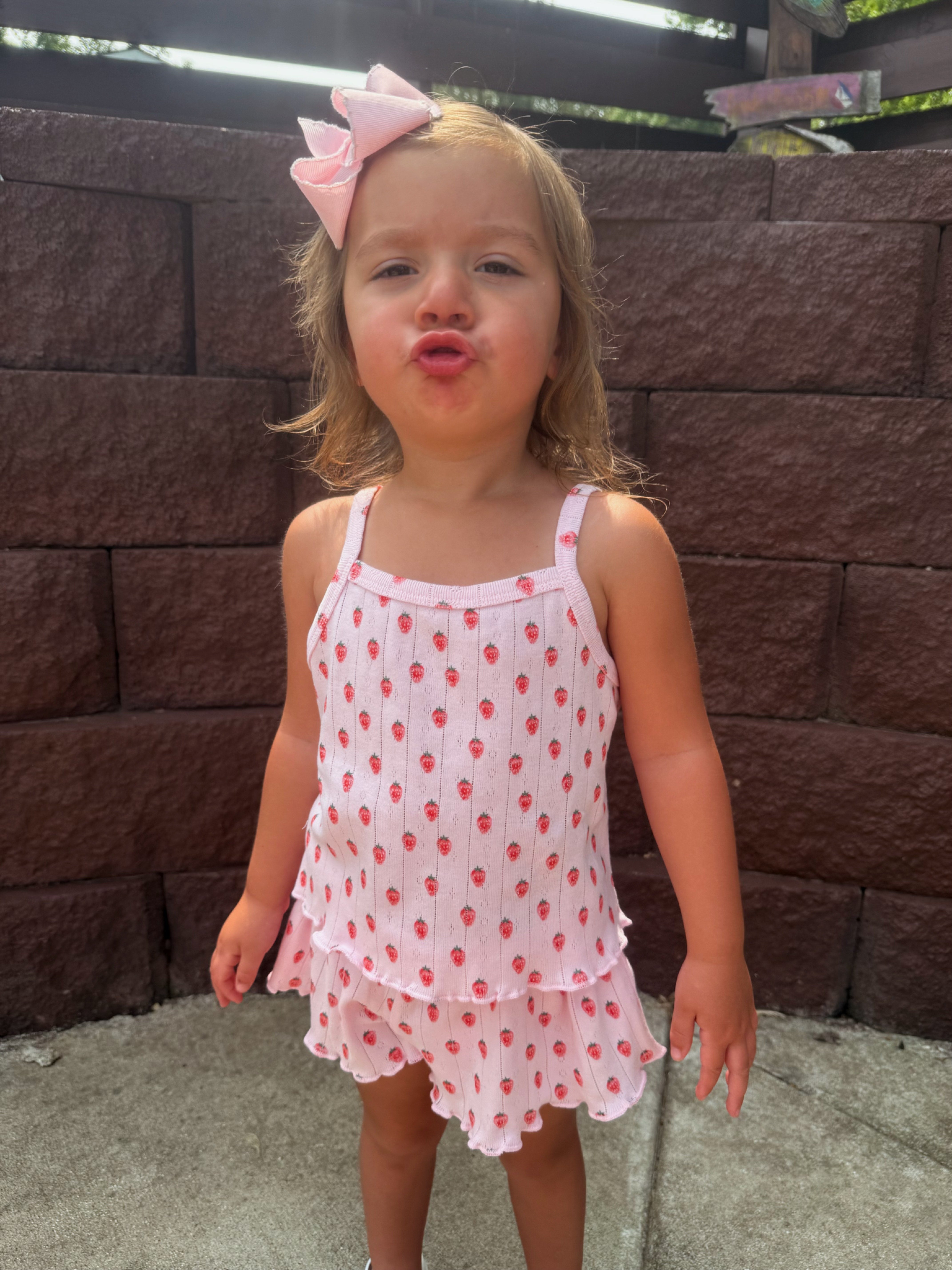 Young girl with a pink bow, wearing a strawberry-patterned outfit, making a playful face outdoors by a stone wall.