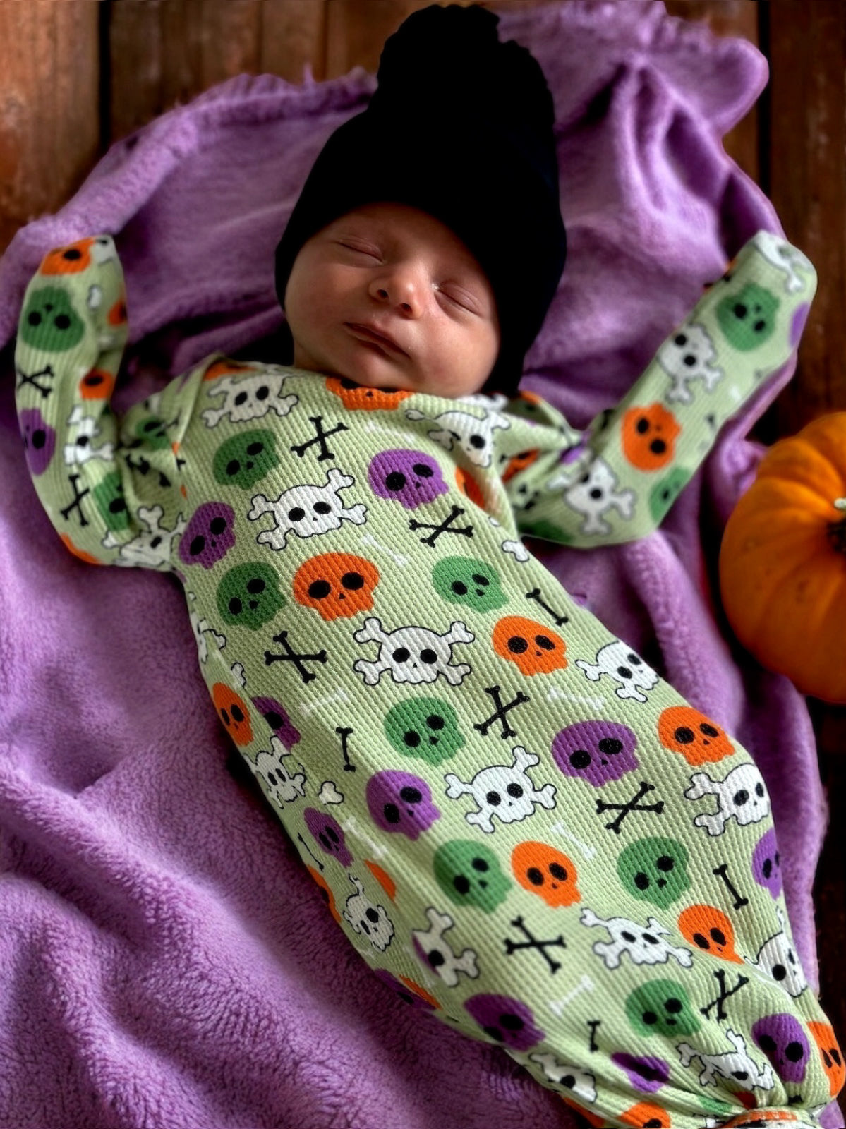 Newborn in a green skull print outfit, resting on a purple blanket next to a small pumpkin, wearing a black beanie.