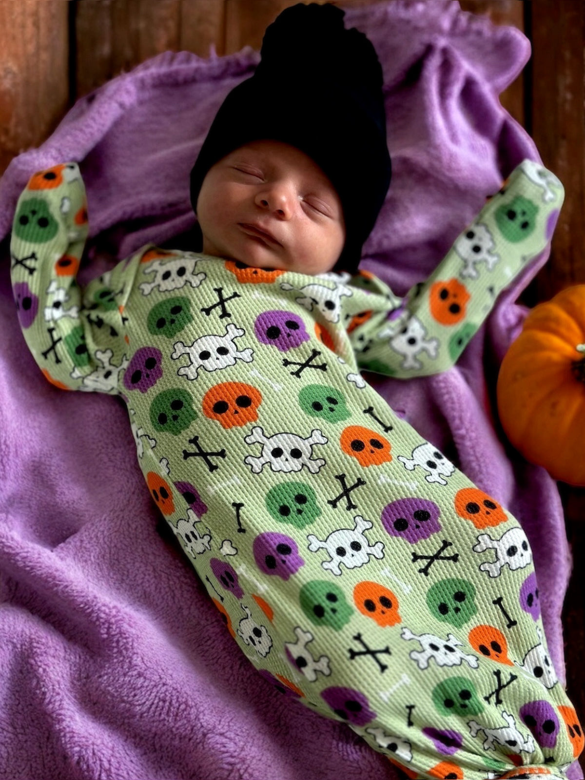 Newborn in a green skull print outfit, resting on a purple blanket next to a small pumpkin, wearing a black beanie.