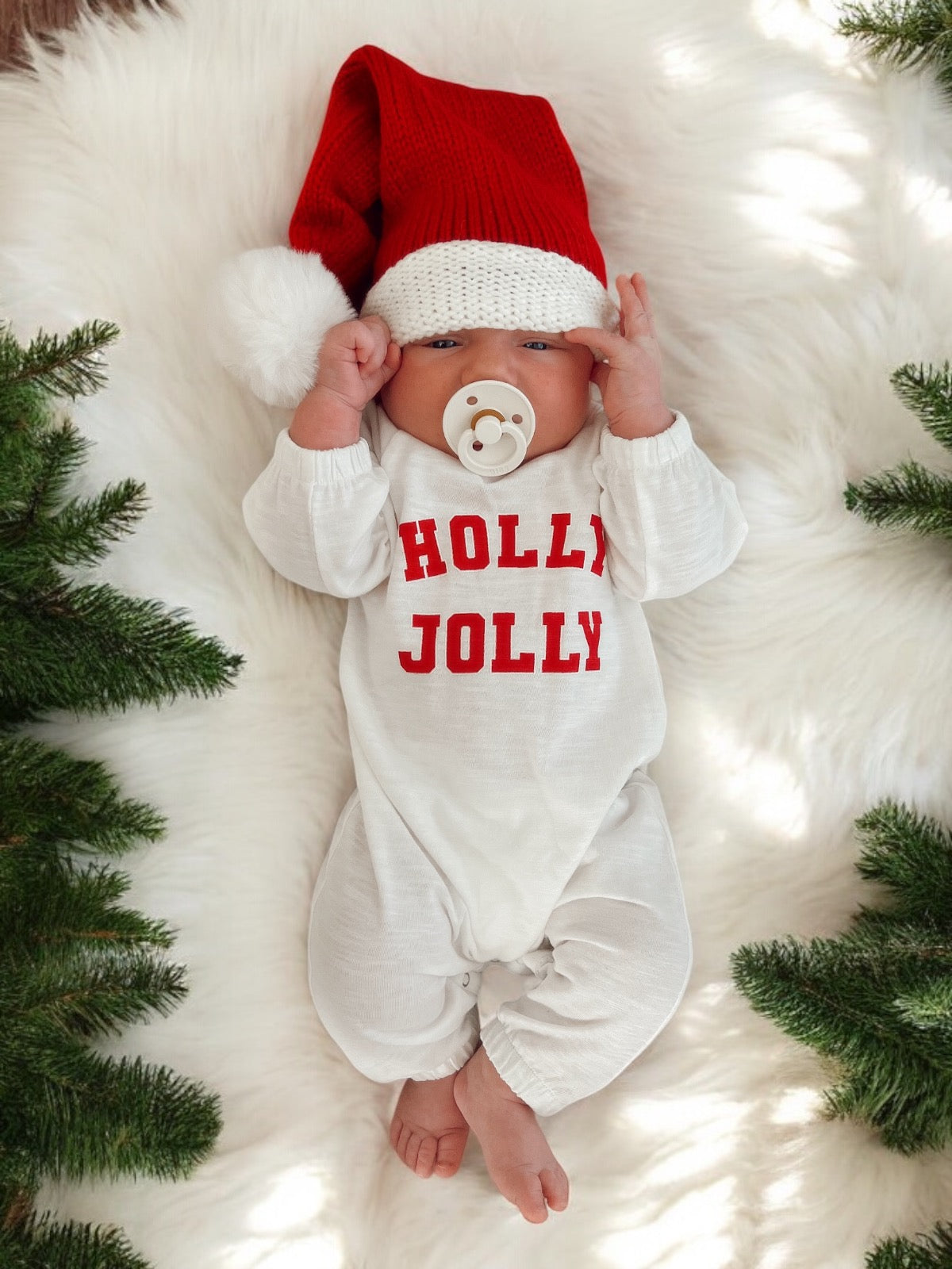Baby in a red and white outfit with a "Holly Jolly" print, wearing a festive Santa hat, surrounded by greenery.