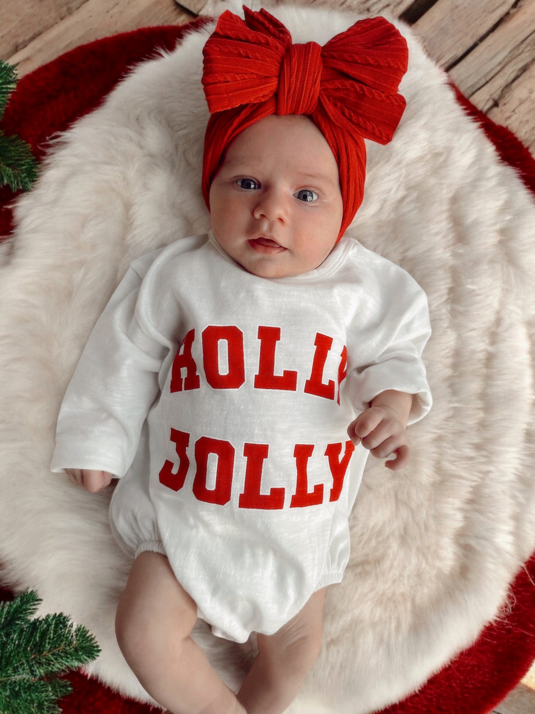 Baby wearing a white "Holly Jolly" outfit with a red bow headband, lying on a fluffy blanket.