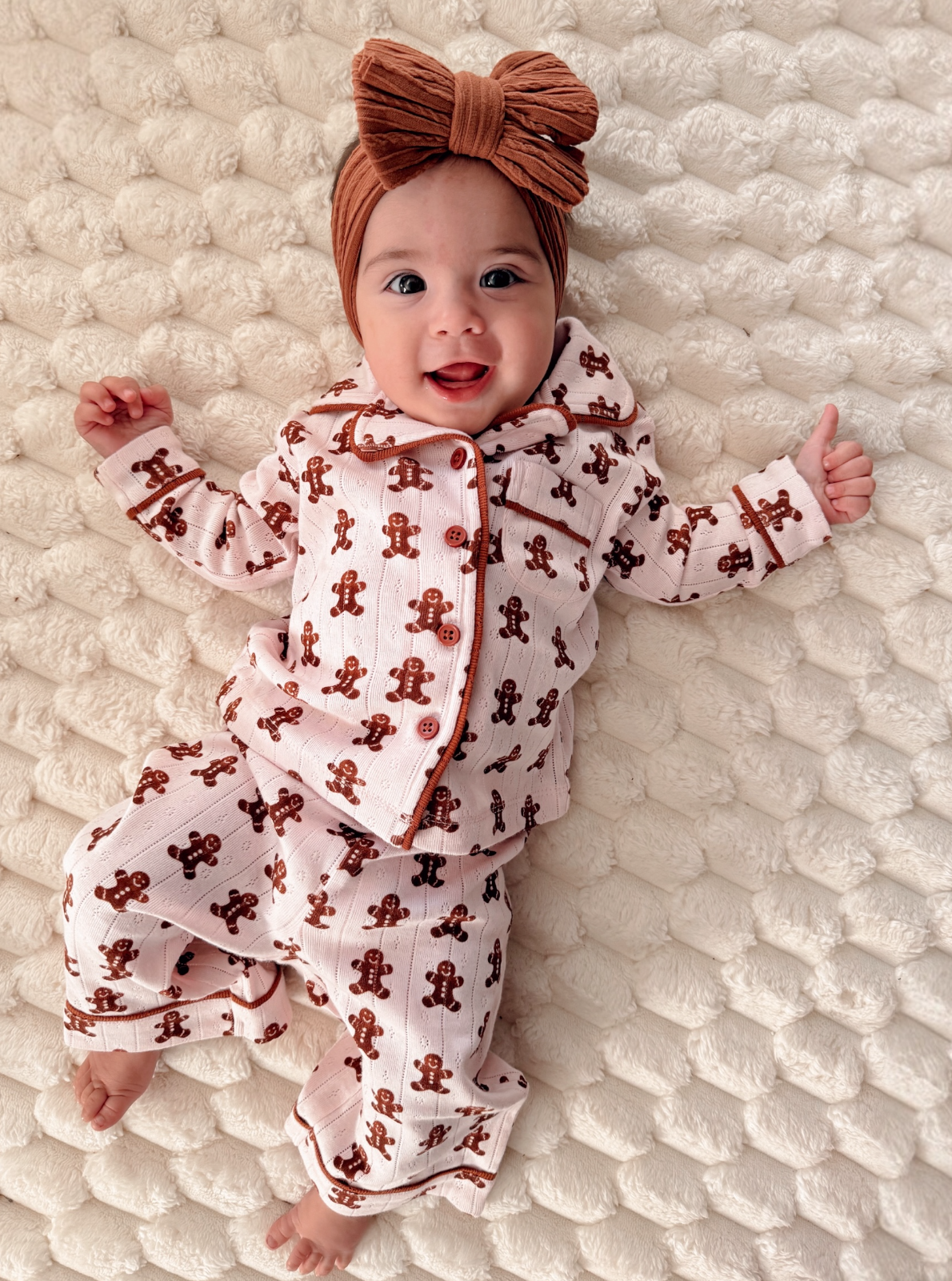 Smiling baby in gingerbread-patterned pajamas on a soft, textured blanket, wearing a large brown headband.