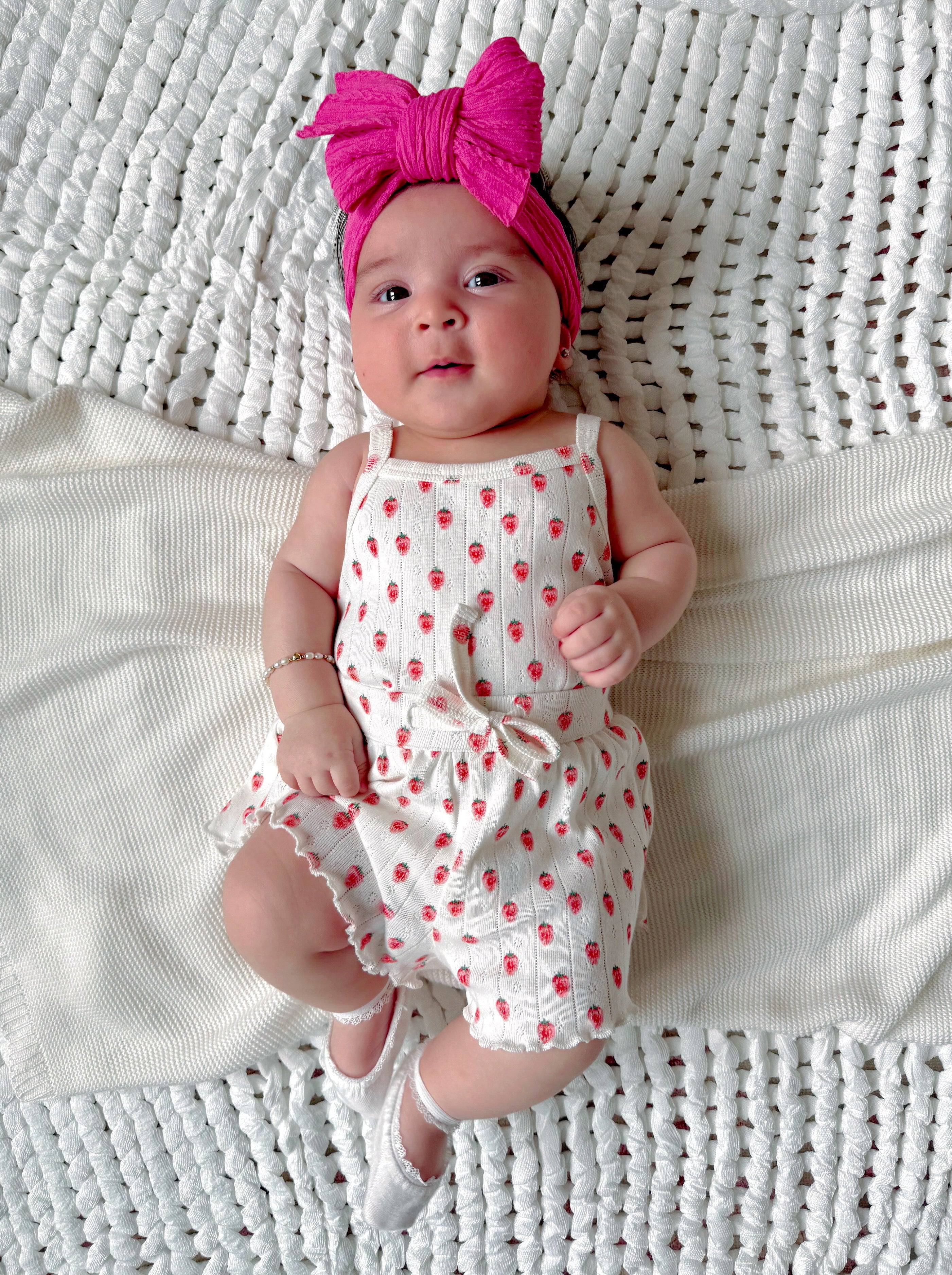 Baby lying on a textured blanket, wearing a white outfit with red strawberries and a pink headband.