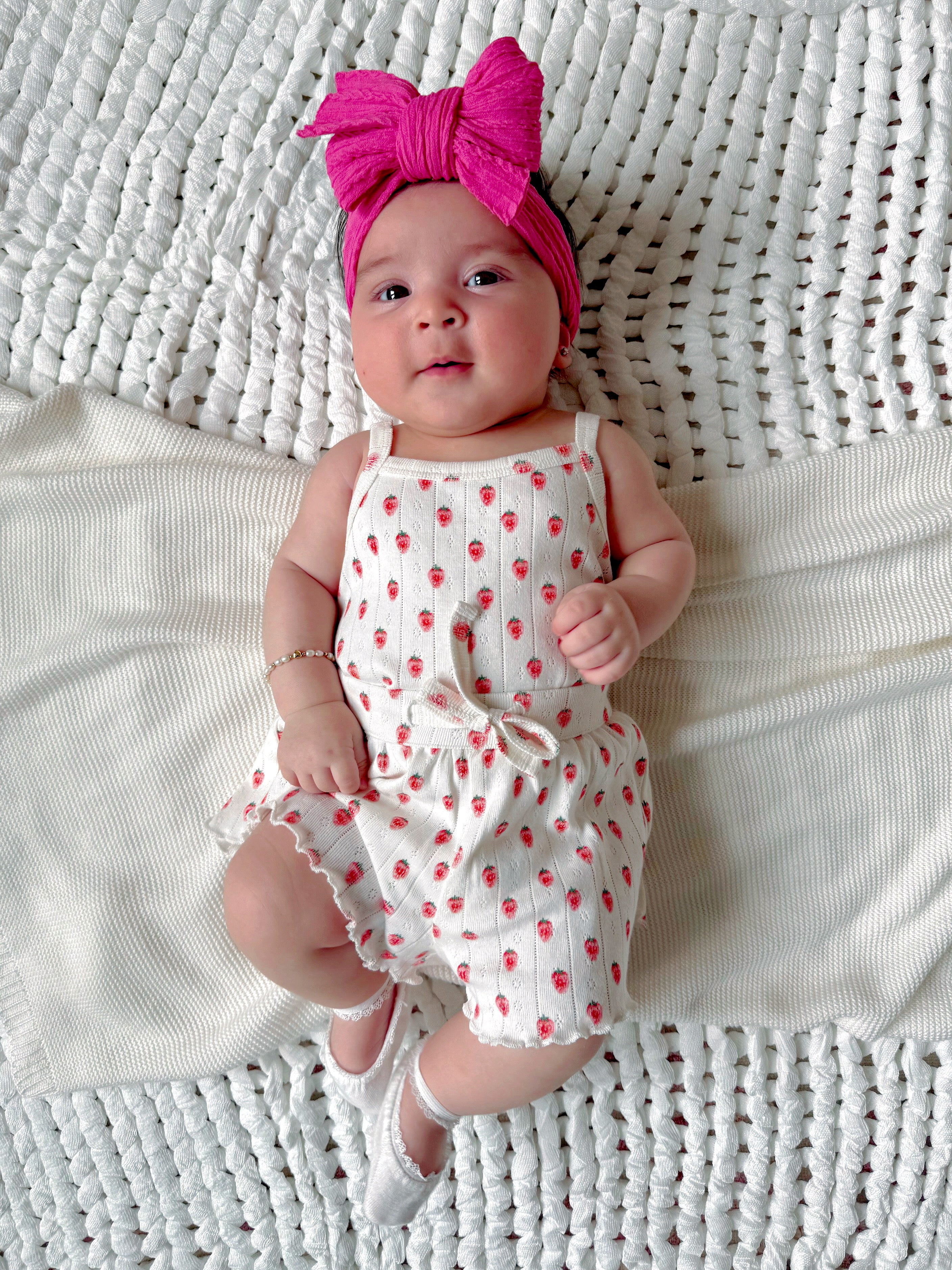 Baby lying on a textured blanket, wearing a white outfit with red strawberries and a pink headband.