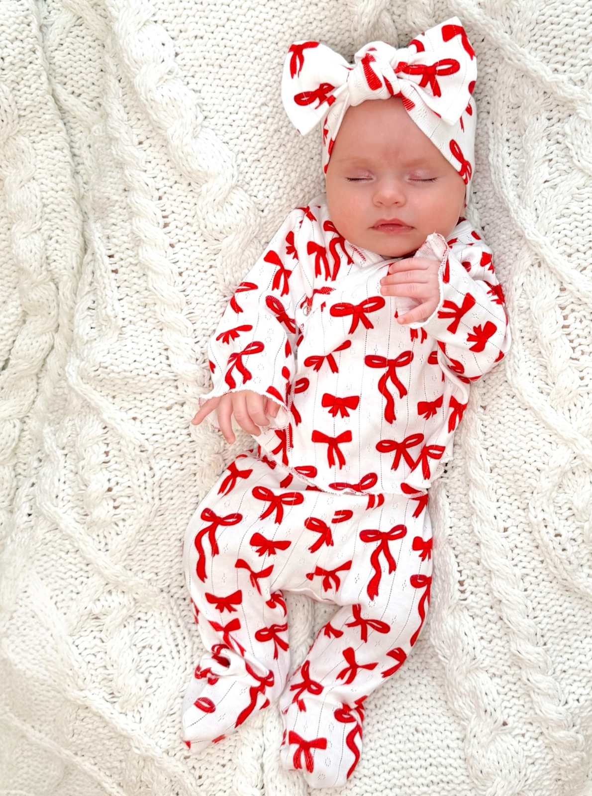 Baby girl in red and white bow-patterned pajama set, resting on a textured white blanket.