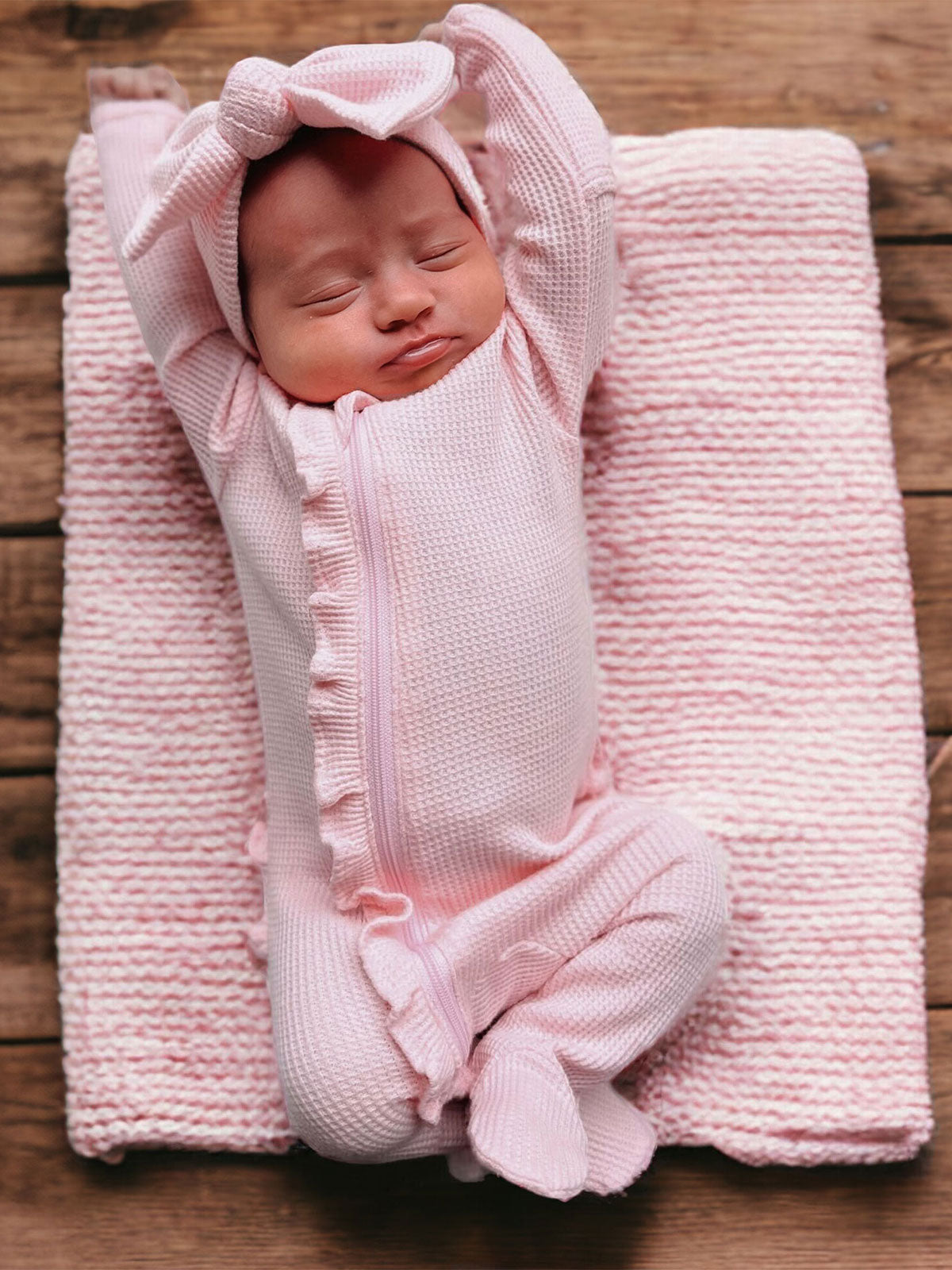 Newborn baby in a pink onesie with a headband, resting on a knitted blanket atop a wooden surface.